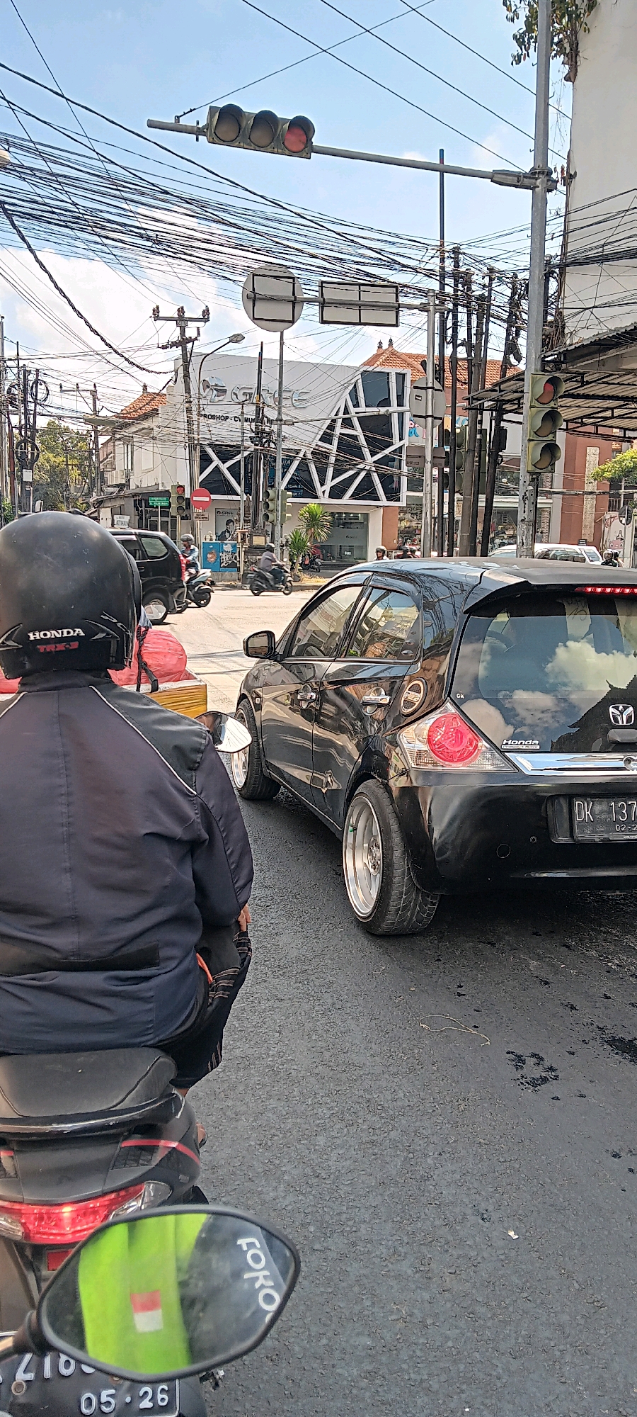 The image depicts a street scene in Denpasar, Indonesia, likely at an intersection, given the presence of traffic lights and multiple vehicles. A person wearing a black helmet and jacket is seated on a black motorcycle, facing away from the camera. The motorcycle has a reflective mirror showing a green vest with the Indonesian flag colors reflected on it, and the word "foko" is visible on the mirror's edge.
In the foreground, a black car with custom silver rims is parked, angled towards the right side of the frame. Its license plate reads "DK 137...". The car's taillights are red and circular. Behind the black car, another black vehicle, possibly an SUV, is also visible. Several other motorcycles and cars are present further down the street, indicating moderate traffic.
The environment is urban, with a large building complex featuring modern architecture in the background. The building has a white facade with geometric patterns and prominent signage that appears to read "GGC" followed by other text. Overhead, a dense network of electrical wires crisscrosses the sky, a common sight in many parts of Indonesia. The sky is clear and blue with some clouds. The road surface is asphalt, showing some wet patches or stains.
The traffic lights in the scene show a red light illuminated on the horizontal traffic signal, suggesting that traffic may be stopped. Another traffic signal on the right shows yellow and red lights illuminated. The overall atmosphere is that of a typical busy street in an Indonesian city during the daytime.