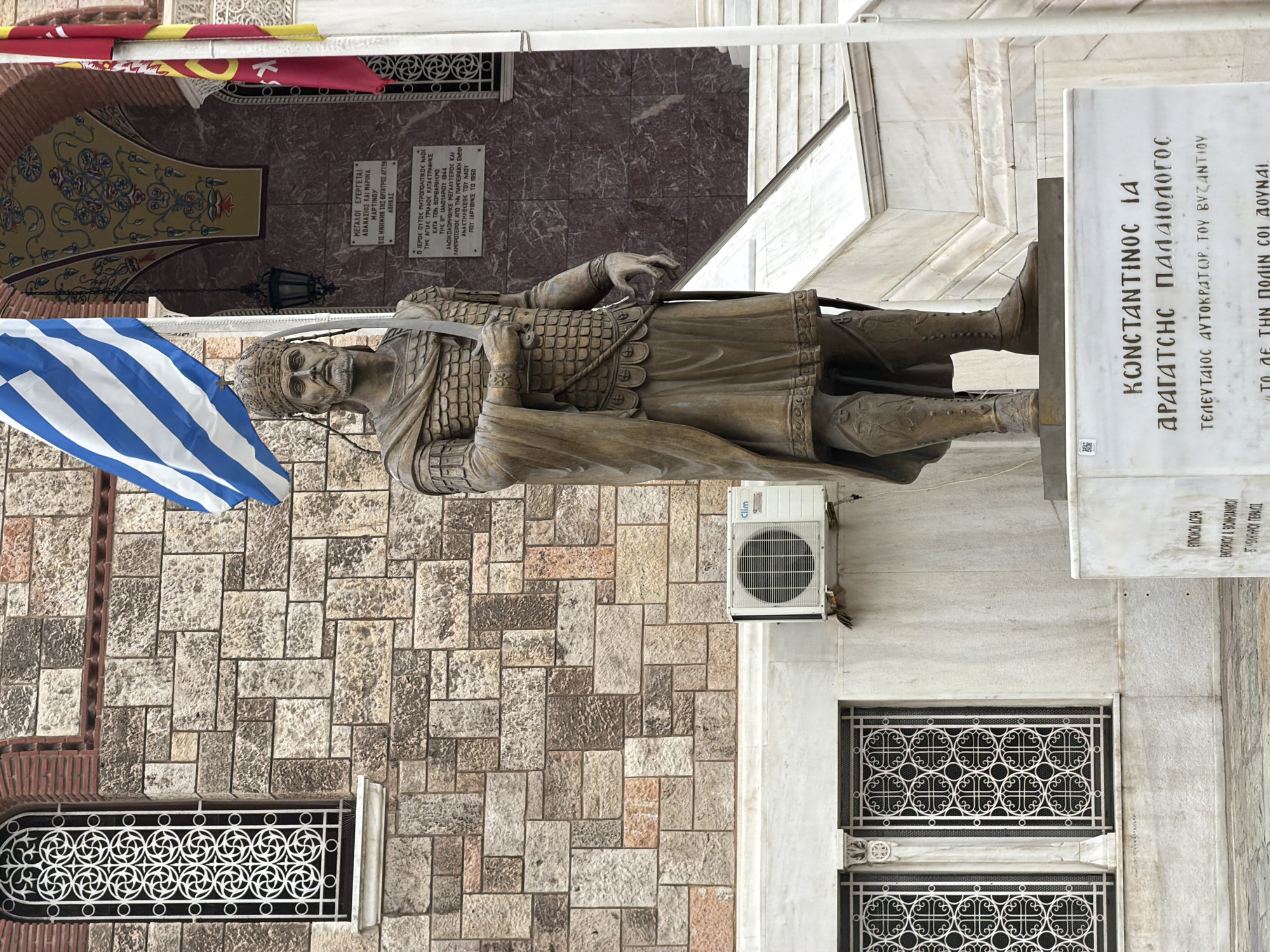 The image depicts a bronze statue of Constantine XI Palaiologos, the last Byzantine Emperor, located in Piraeus, Greece. The statue stands on a marble pedestal with inscribed Greek text. A Greek flag is positioned to the left of the statue. The backdrop includes a stone wall with a window featuring a decorative metal grille, and a wall-mounted air conditioning unit. A marble plaque with Greek text is above the statue. Red and yellow flags are hung on the building in the background.