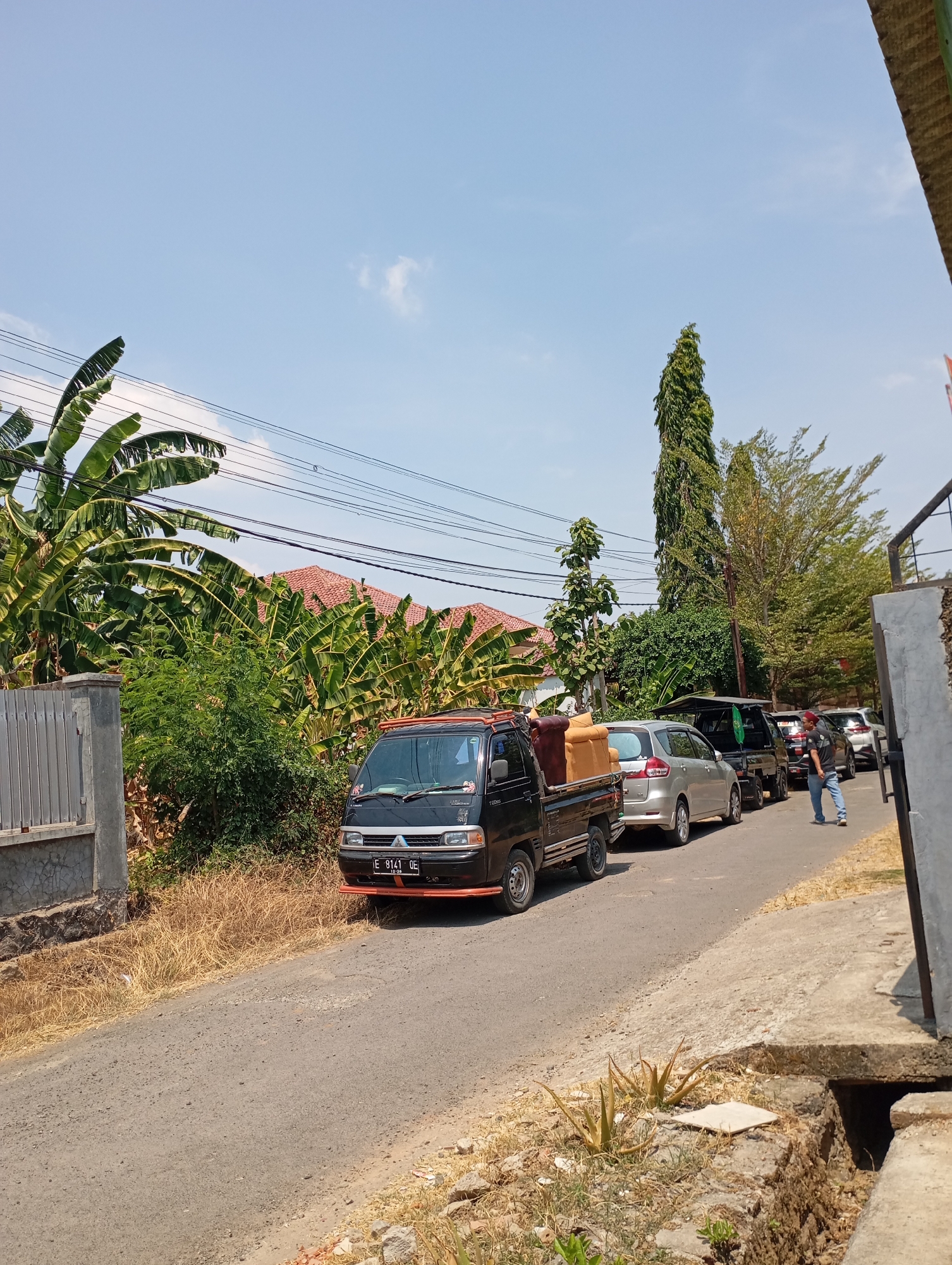 A black pickup truck carrying furniture is parked on a narrow, paved road in an outdoor environment. The truck has a license plate that reads "E 8141 QE". To the left of the truck is a grey metal gate and fence, with lush green banana trees behind it. Further back, red-tiled roofs of houses are visible amongst dense foliage. The road is lined with dry grass and some greenery. In the distance, a tall, thin tree stands out against a clear blue sky with a few scattered clouds. Other vehicles, including a silver car and a dark SUV, are also present on the road, suggesting a traffic queue. A person wearing a blue shirt and jeans is walking away from the camera on the right side of the road. The scene appears to be in a rural or suburban area, likely in Indonesia, given the style of the houses and vehicles.