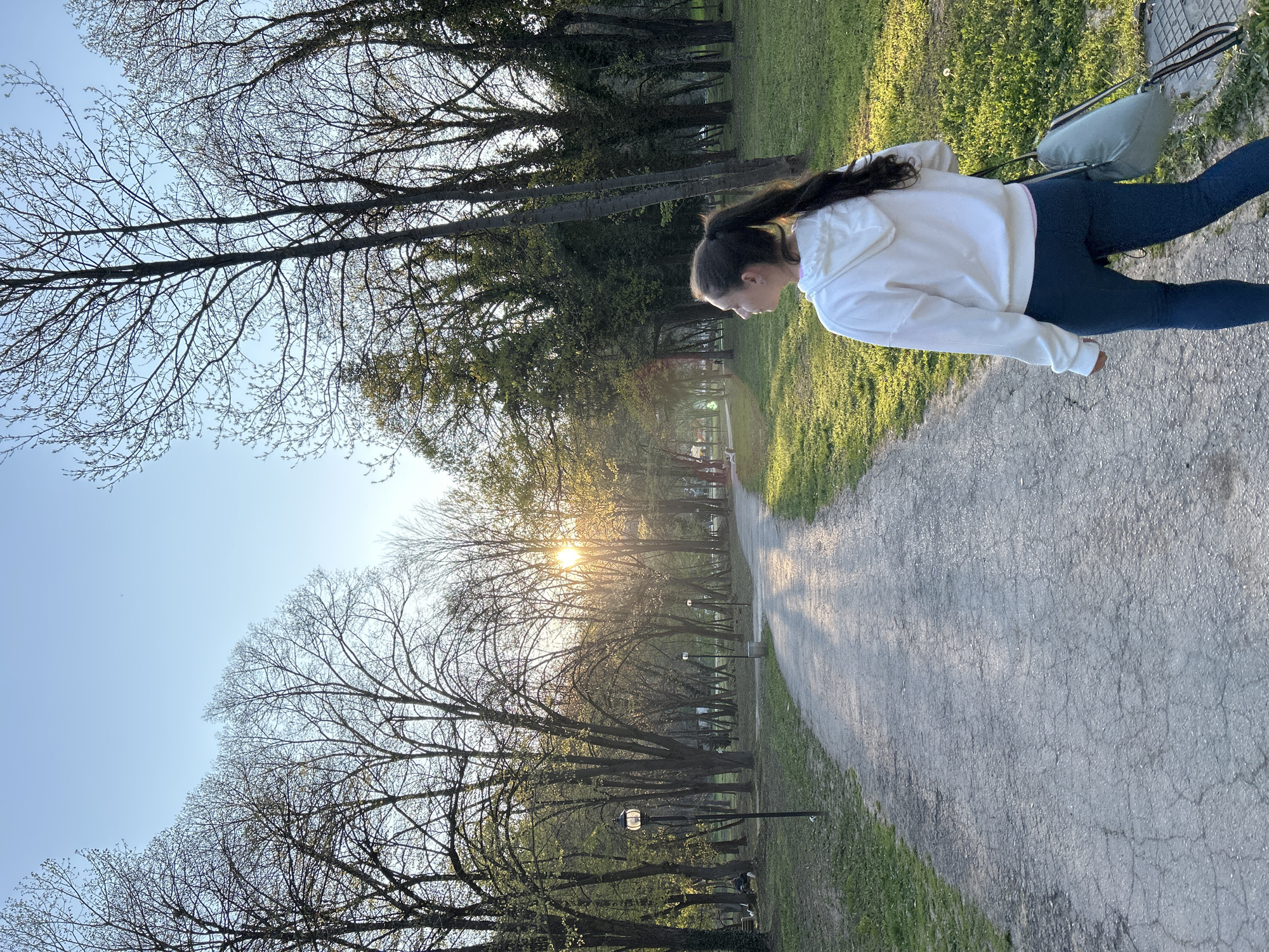 A young woman is walking on a paved path in a park at sunset. The path is flanked by trees, some without leaves, silhouetted against the sun. The woman is wearing a white hooded sweatshirt and blue leggings, and she has a light blue purse on her shoulder. The sky is pale blue near the horizon. The park appears to be in Pleven, Bulgaria.
