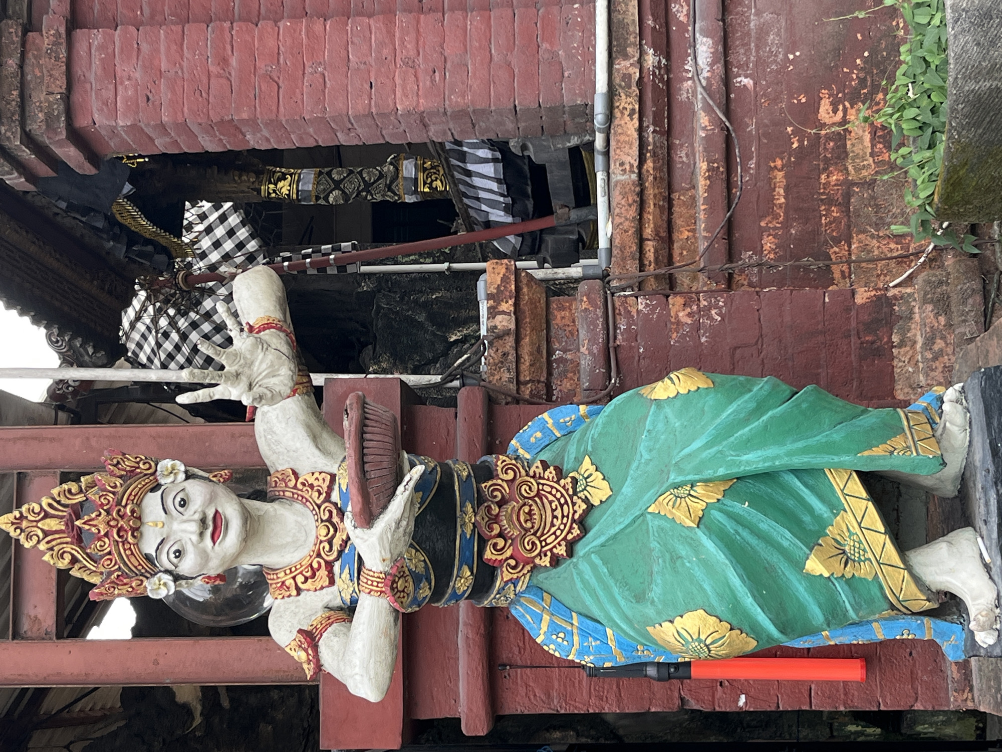 A full-body, outdoor, eye-level shot captures a vibrant, ornate statue of a Balinese dancer in Kuta, Indonesia. The statue, painted in bright colors, stands before a weathered red brick wall and a traditional Balinese temple structure adorned with black and white checkered cloth. The dancer wears a golden headdress, a beaded chest piece, and a flowing green sarong decorated with gold floral patterns. Her hands are held forward, one carrying a small red bowl. Vines and greenery grow in the foreground. The statue's face is painted white with red lips and dramatic eye makeup. The red brick wall shows signs of age and peeling paint.