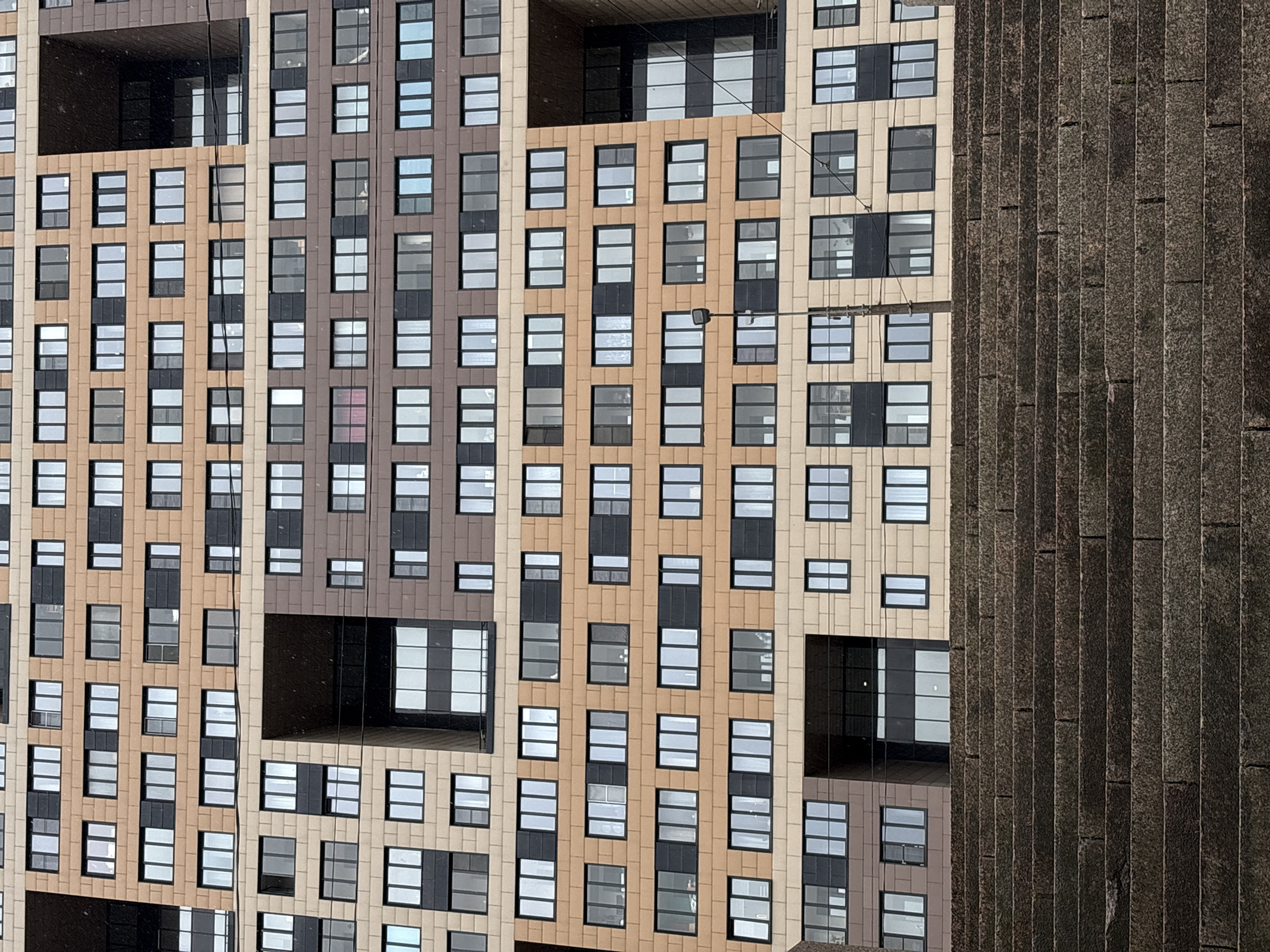 The image shows the exterior of a modern building in Saint Petersburg, Russia. The building has a geometric facade with a pattern of windows and panels in varying shades of brown and beige. To the right of the building, there's a textured concrete structure with vertical lines. There are no people visible in the frame. The lighting suggests an overcast day. Electrical lines are visible in the sky.