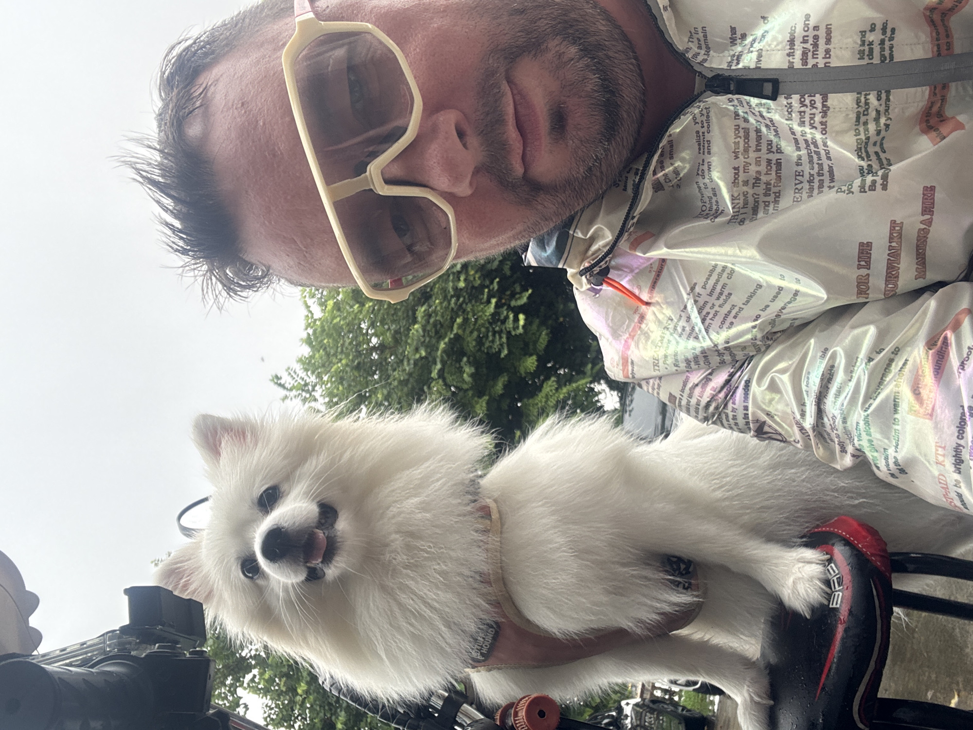 A man and a white Pomeranian dog are in a selfie outdoors. The man is wearing oversized, light-colored sunglasses and a patterned jacket with text on it. The dog is sitting on a black seat with red trim, possibly a motorcycle seat. The background shows greenery and an overcast sky. The image appears to be taken in a semi-urban or suburban environment, possibly on a street in Nanhai District, China. The man has short, dark hair and a beard. The dog has its mouth slightly open and is looking at the camera.