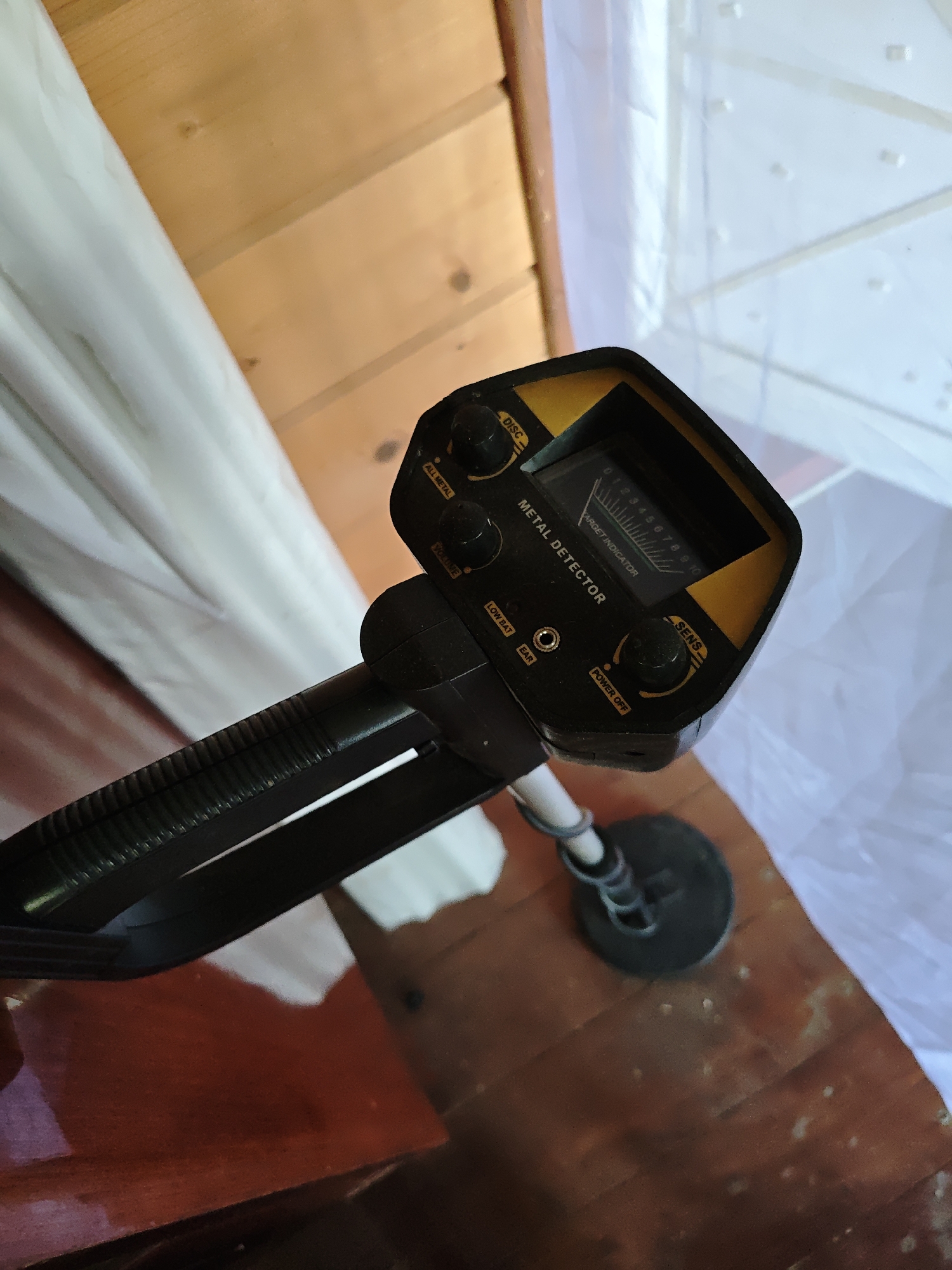 A metal detector sits indoors on a wooden floor, positioned next to a wooden wall and a semi-transparent white curtain. The metal detector is black with yellow accents and features a control panel with dials and an indicator screen. The surrounding environment suggests an indoor setting with natural light, possibly a residential or storage area. The location of Боровково, Russia, is not evident from the image.
