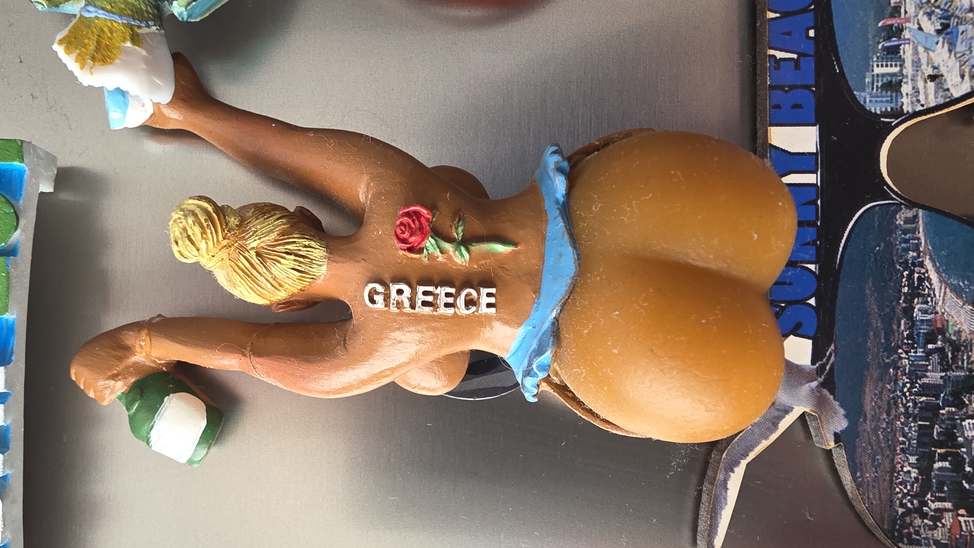 A close-up shot displays a magnet depicting a woman lying prone with a large, stylized buttocks. The woman is painted in a light brown color with a small rose on her chest and the word "GREECE" in white letters nearby. She has blonde hair styled in a bun. The background includes a partially visible postcard with the words "SUNNY BEACH." The location and city of Blagoevgrad, Bulgaria are not relevant to the image as depicted.