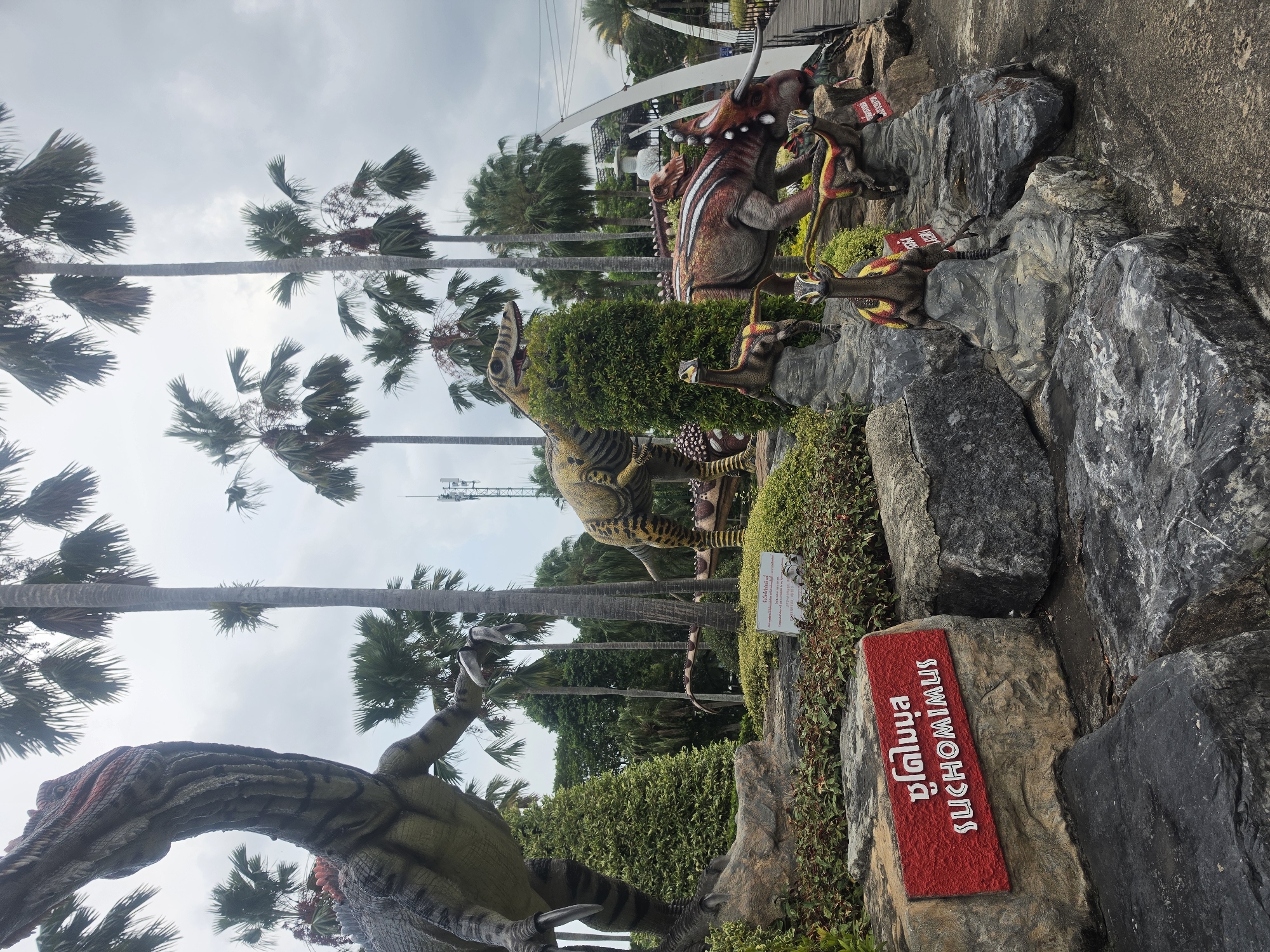 The image depicts a dinosaur display in Na Chom Thian, Thailand. Multiple static dinosaur models are arranged amidst rocks, shrubbery, and palm trees. A red sign with white Thai and English text reads "Suchomimus." The sky is overcast. A white pedestrian bridge is visible in the background. A cellular tower is also visible. The image orientation is rotated approximately 90 degrees counter-clockwise.