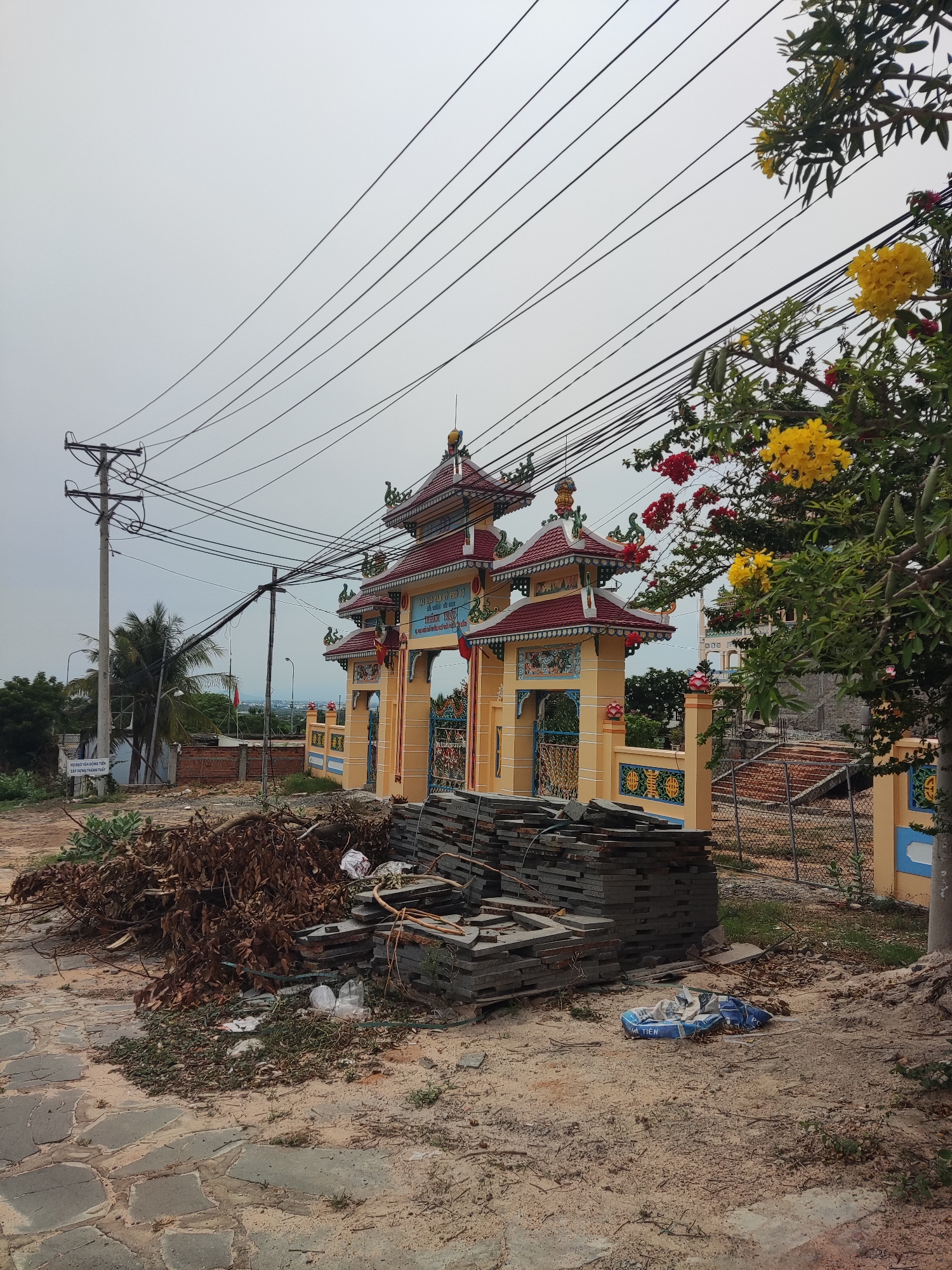 The image depicts a religious building, likely a temple or shrine, in Unknown, Unknown. The building is painted in yellow and red with ornate architectural details, including a multi-tiered roof. A stack of stone tiles lies in front of the building, along with debris such as dried leaves and plastic bags. A utility pole with multiple wires stands to the left of the building. A tree with yellow and red flowers is on the right side of the frame. The ground is dirt and stone paving.