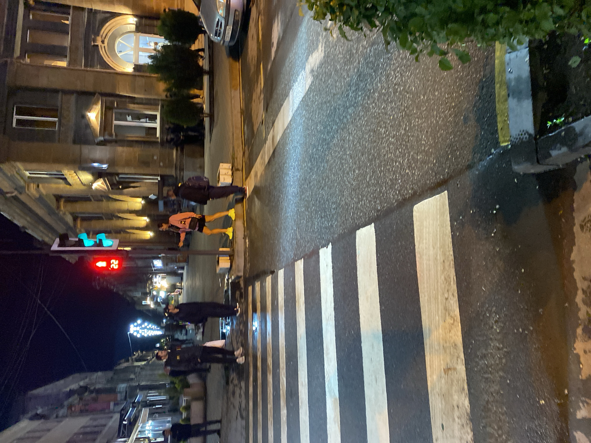 The image depicts a nighttime street scene in Gyumri, Armenia. A pedestrian crossing is visible in the foreground, with wet asphalt indicating recent rain or moisture. In the background, a traffic light shows a red signal, with the number 22 displayed on a digital counter beneath it, suggesting a countdown or traffic information. Several people are present on the sidewalk and street, some appearing to wait at the intersection. Buildings with illuminated windows line the street, and streetlights are also visible, casting a glow on the surroundings. A car is parked on the right side of the image. The overall atmosphere is that of a typical urban environment at night, with elements of traffic control and pedestrian activity.