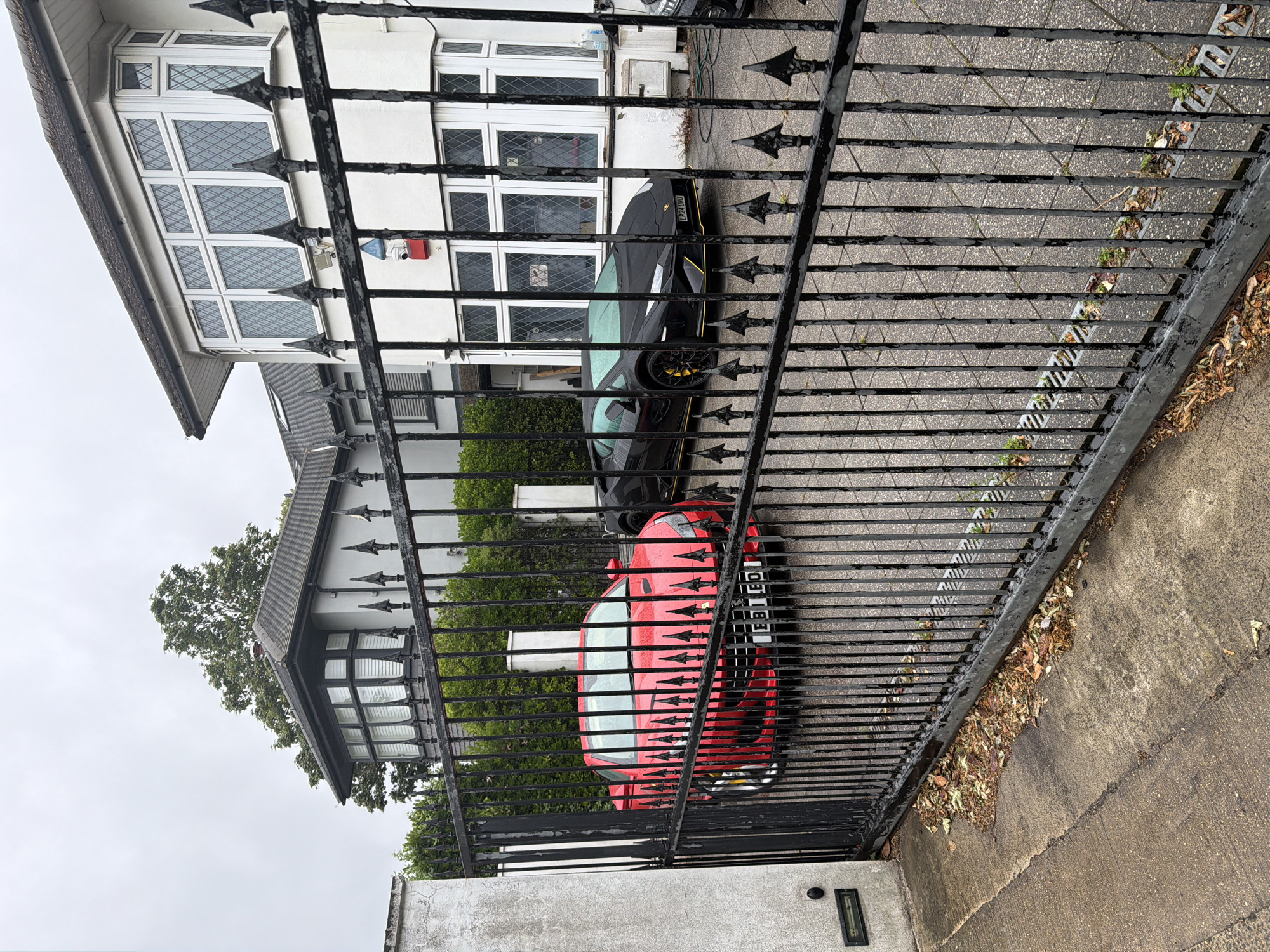 A black wrought-iron gate stands in the foreground of the image, with tall, pointed finials adorning its upper edge. Beyond the gate, a driveway is visible, wet from rain. On the left side of the driveway is a bright red sports car, a Ferrari, with its license plate reading "EB11 EQ". To the right of the red car is a black Lamborghini sports car with yellow accents on its trim. The cars are parked in front of a large, modern house. The house features a distinctive bay window with diamond-shaped panes on the upper floor and a white facade. A security camera and a red alarm box are mounted on the exterior wall of the house. The sky is overcast, suggesting a cloudy day. The environment is a residential area, likely in a city, given the architecture of the house and the presence of multiple high-end vehicles. The presence of fallen leaves on the ground suggests it may be autumn. The image appears to have been taken in London, United Kingdom, given the license plate format of the vehicles.