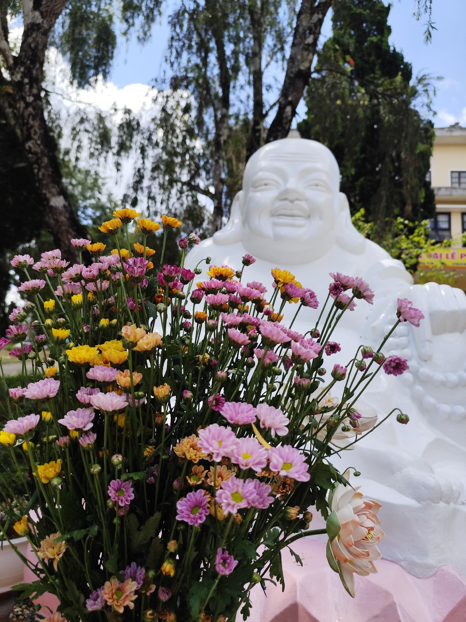 The image shows a white, smiling Buddha statue partially obscured by a vibrant bouquet of flowers. The flowers are predominantly yellow, pink, and light orange, arranged in a dense cluster. One larger, pale pink artificial lotus blossom is visible at the lower right. The statue sits outdoors, with trees visible in the blurred background. The context suggests this is located in Da Lat, Vietnam, given previous context. A building with windows is partly seen behind the statue.