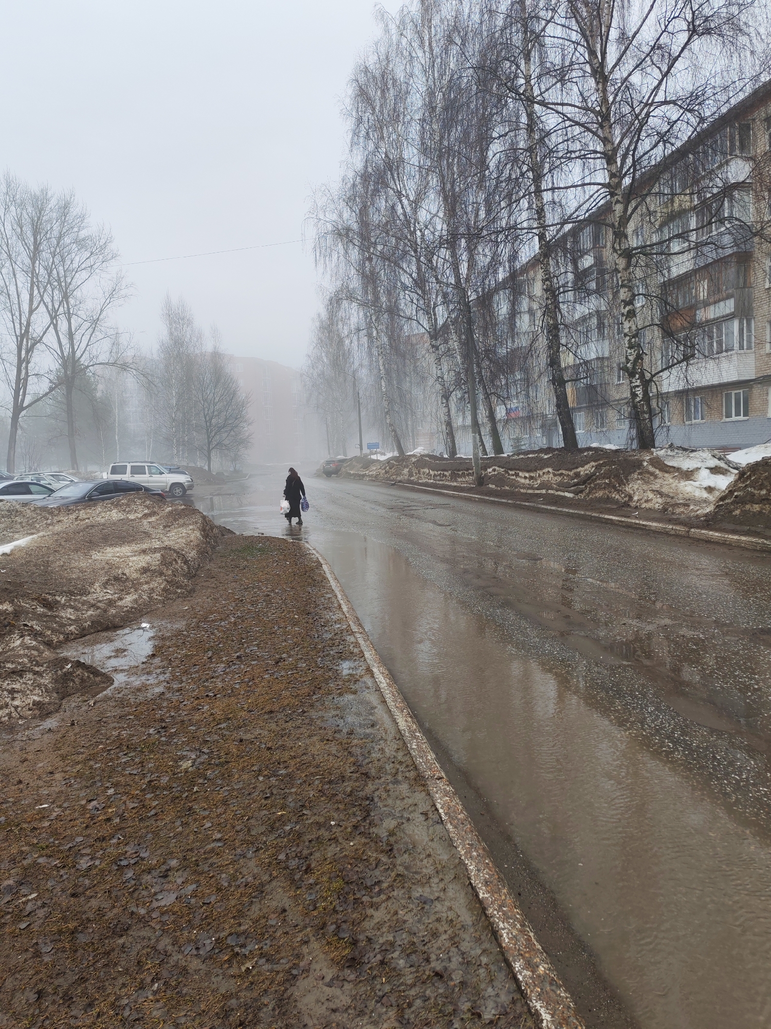 The image depicts a street in Chaikovsky, Russia on a foggy, overcast day. The road is wet with standing water, reflecting the sky and surrounding buildings. On the right side of the road is a multi-story residential building, and bare trees line the street. Piles of dirty snow and mud are heaped on both sides of the street, indicating recent snow melt. A person is walking along the side of the road, carrying bags, presumably heading towards or from the building. Several parked cars are visible on the left side of the image. The overall atmosphere is bleak and damp.