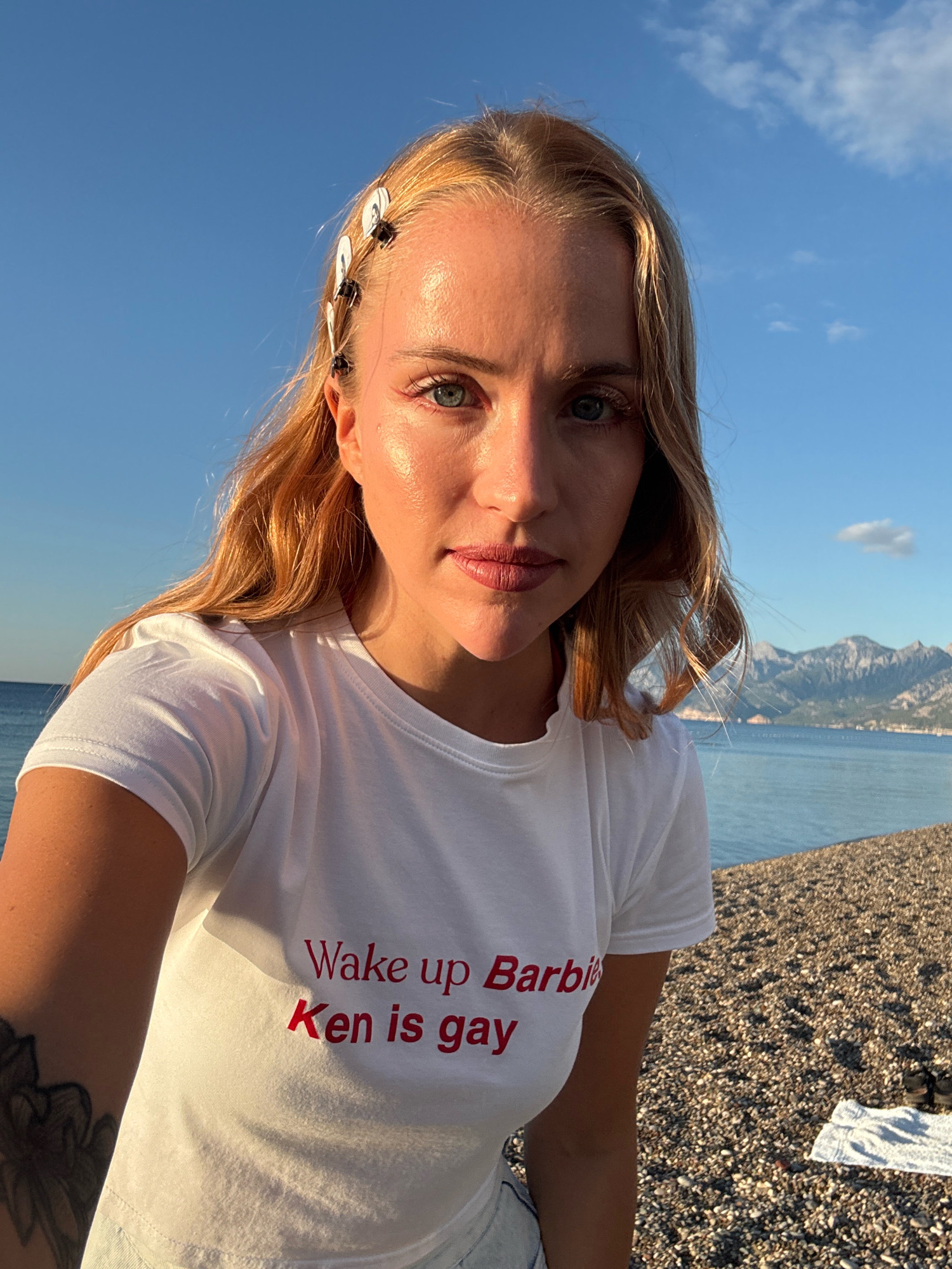 A woman with blonde hair, wearing a white t-shirt with red text that reads "Wake up Barbie, Ken is gay", stands on a pebbled beach. The sea is visible behind her, with mountains in the distance. The sky is blue with a few wispy clouds. The scene appears to be set during a sunny day, possibly at sunset given the warm lighting. The location is the city of Antalya, Turkey.