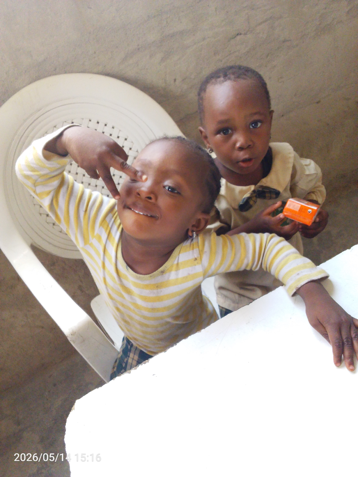 Two children are captured in an indoor setting, likely in Lokoja, Nigeria, as per the instructions. The child in the foreground, seated on a white plastic chair, wears a yellow and white striped long-sleeved shirt and what appears to be a plaid bottom. They are making a gesture with their right hand near their eye, and their left arm rests on a white table. The child in the background, partially obscured, is wearing a neutral-toned button-down shirt and holding a small orange object. The walls are light colored, suggesting a simple interior. The date stamp visible in the lower left corner is "2026/05/14 15:16".
