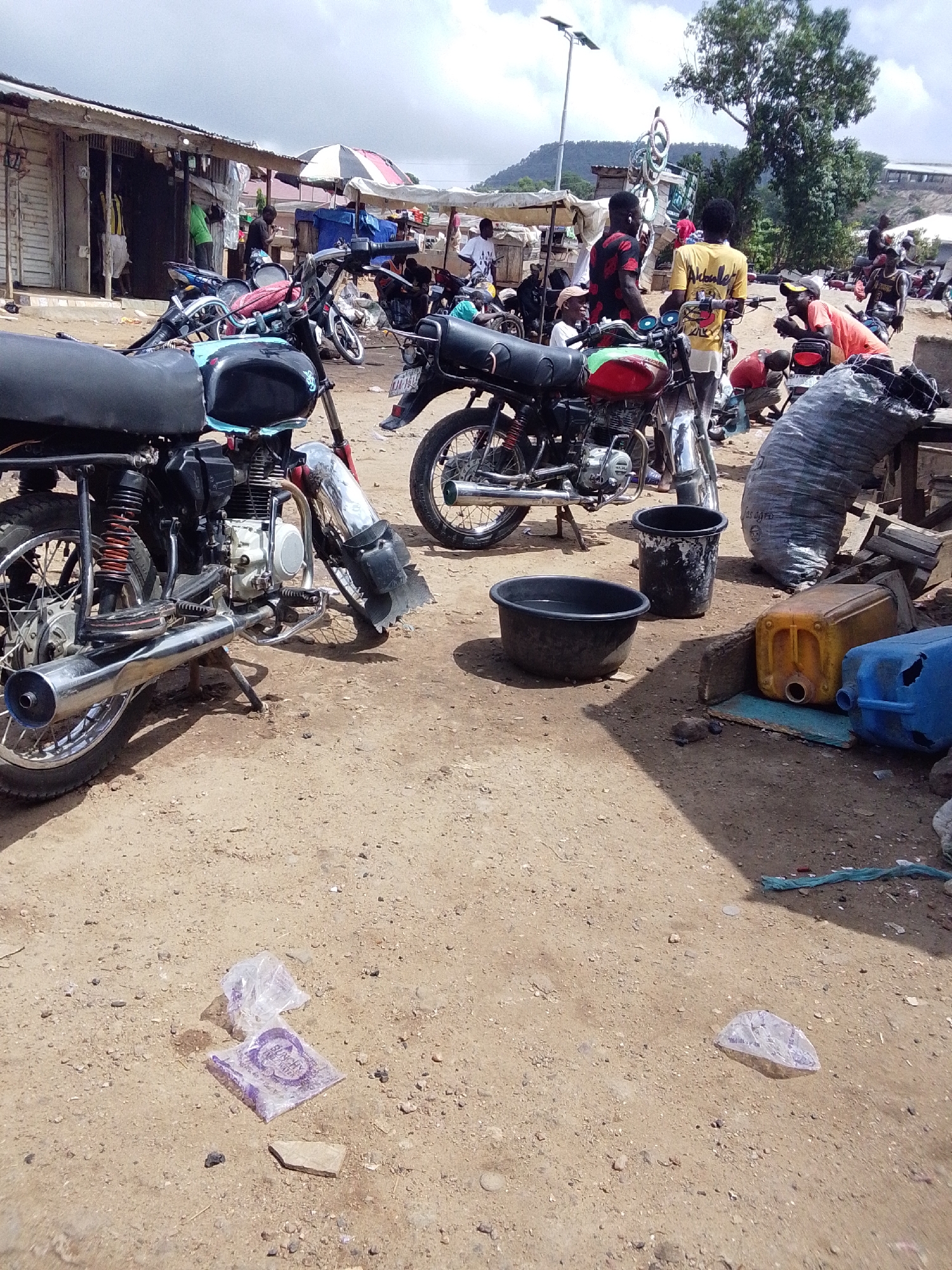 The image depicts an open-air area in Lokoja, Nigeria. Several motorcycles are parked on a dirt ground, with evidence of commercial activity or repairs taking place. There are people present, some standing and interacting. A large sack and several plastic containers are visible. Additionally, some waste materials, possibly packaging, are scattered on the ground. The structures in the background suggest an informal market or roadside business area.