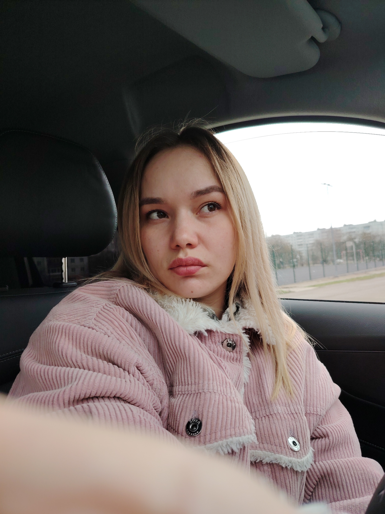 A young woman with long, blonde hair is depicted in a close-up, half-body shot, sitting in the passenger seat of a car. She is wearing a soft pink, corduroy jacket with white, fluffy trim around the collar, cuffs, and button placket. Her head is turned to her right, and she is looking out the car window with a somewhat pensive or contemplative expression. Her lips are slightly parted, and her eyes are directed towards something outside the frame.
The car's interior is visible, with a dark headrest to her left and the window on her right showing an overcast sky and a distant urban landscape. Outside the window, there are visible buildings, bare trees, and what appears to be a field or open space, typical of a city like Chaikovsky, Russia, during cooler weather. The light suggests it is daytime, possibly early morning or late afternoon, given the diffused light from the cloudy sky.
Notable details include the texture of her corduroy jacket and the white trim. On her jacket's snap buttons, the text "SOVA ORIGINAL" is visible, along with some smaller, less discernible text that appears to be "FABRICS". The car's sun visor is partially visible at the top of the frame, indicating the interior environment. Her left hand or arm is partially in the bottom left foreground, out of focus. The overall mood is quiet and observational.
