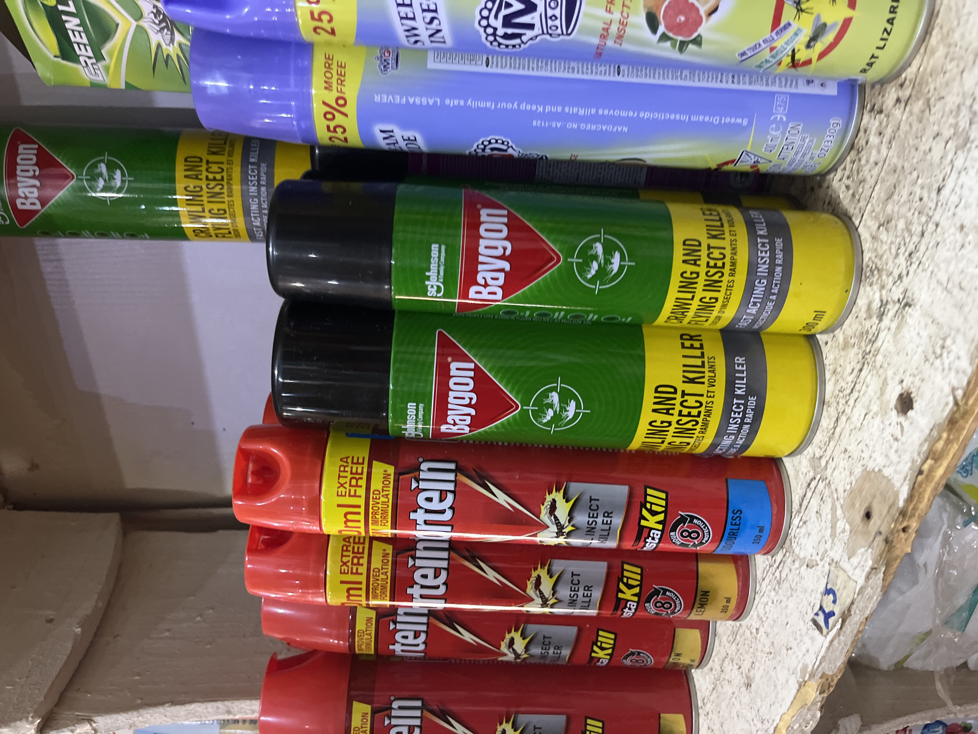 The image shows a shelf in a store, likely a pharmacy or general goods store, in Bukuru, Nigeria, displaying various aerosol cans of insect repellent. The cans are arranged in rows, with some in the foreground and others further back. The products visible include "Baygon" insect killer in green cans, "InstaKill" in red cans, and other brands in purple and yellow cans, some of which indicate "25% More Free." The shelf itself appears to be made of aged wood with visible wear and tear. In the background, other shelves and products are partially visible, suggesting a retail environment. No people are present in the immediate frame of the image. The lighting appears to be artificial, typical of an indoor retail space.