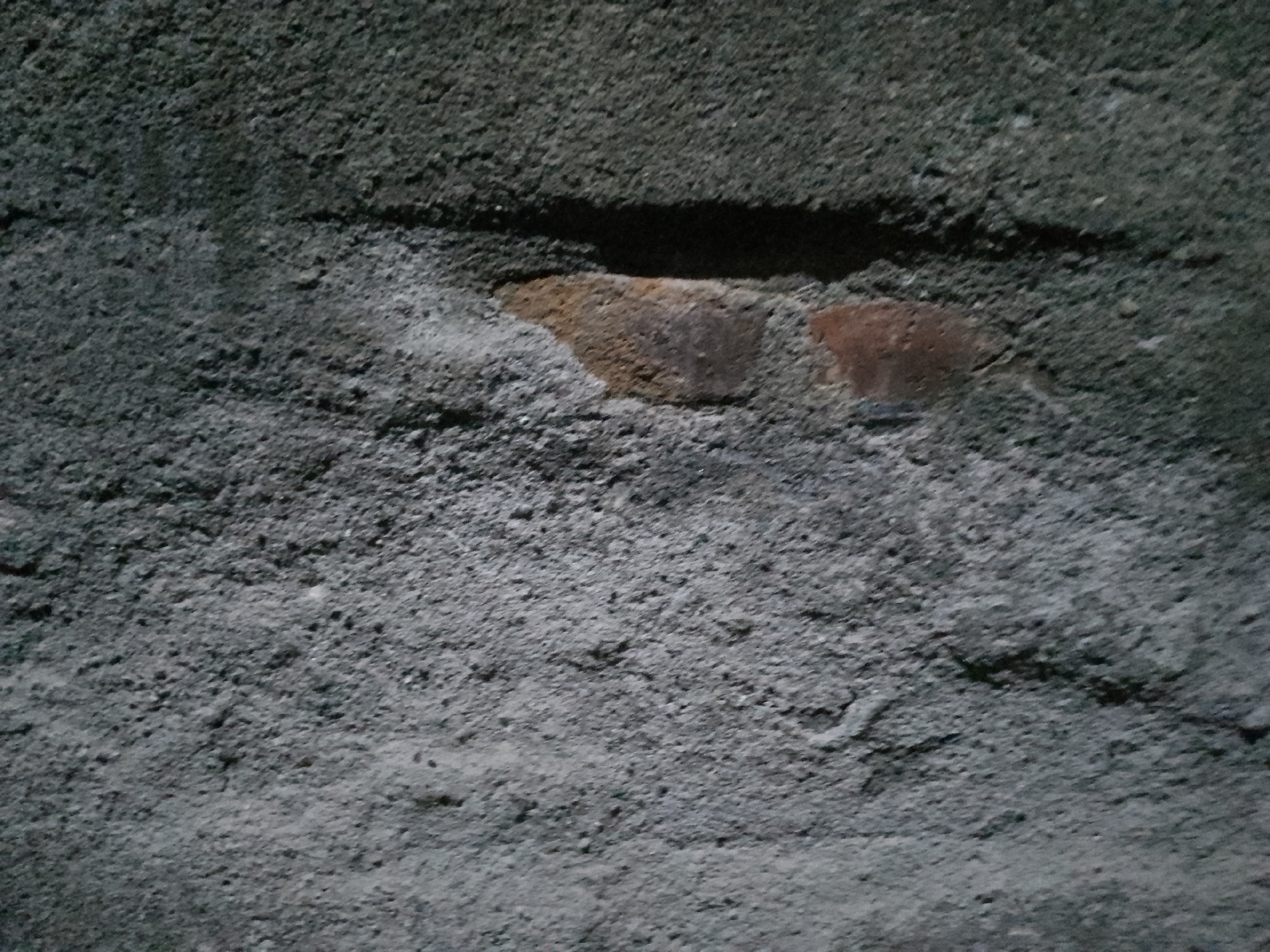 The image shows a close-up view of a weathered, textured wall with visible brickwork peeking through the plaster. The wall is predominantly gray, with rough, uneven surfaces indicating age and exposure to the elements. In the central part of the image, a section of reddish-brown brick is exposed, suggesting that the wall is likely made of brick that has been plastered over. There are no people or animals visible in the image, and the environment appears to be outdoors, given the texture and condition of the wall. The lighting is dim, casting shadows that accentuate the texture of the surface. The location is specified as the city of Kauman, Indonesia.