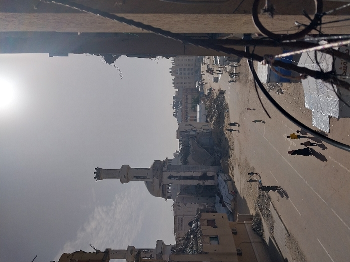 The scene depicts a street with damaged buildings, including a mosque with a minaret. Rubble and debris are visible. Several people are walking along the street, and there appear to be aid tents set up. The sky is overcast, and the sun is visible. Overhead, wires and a balcony railing are in view. The overall impression is of destruction and the aftermath of conflict.