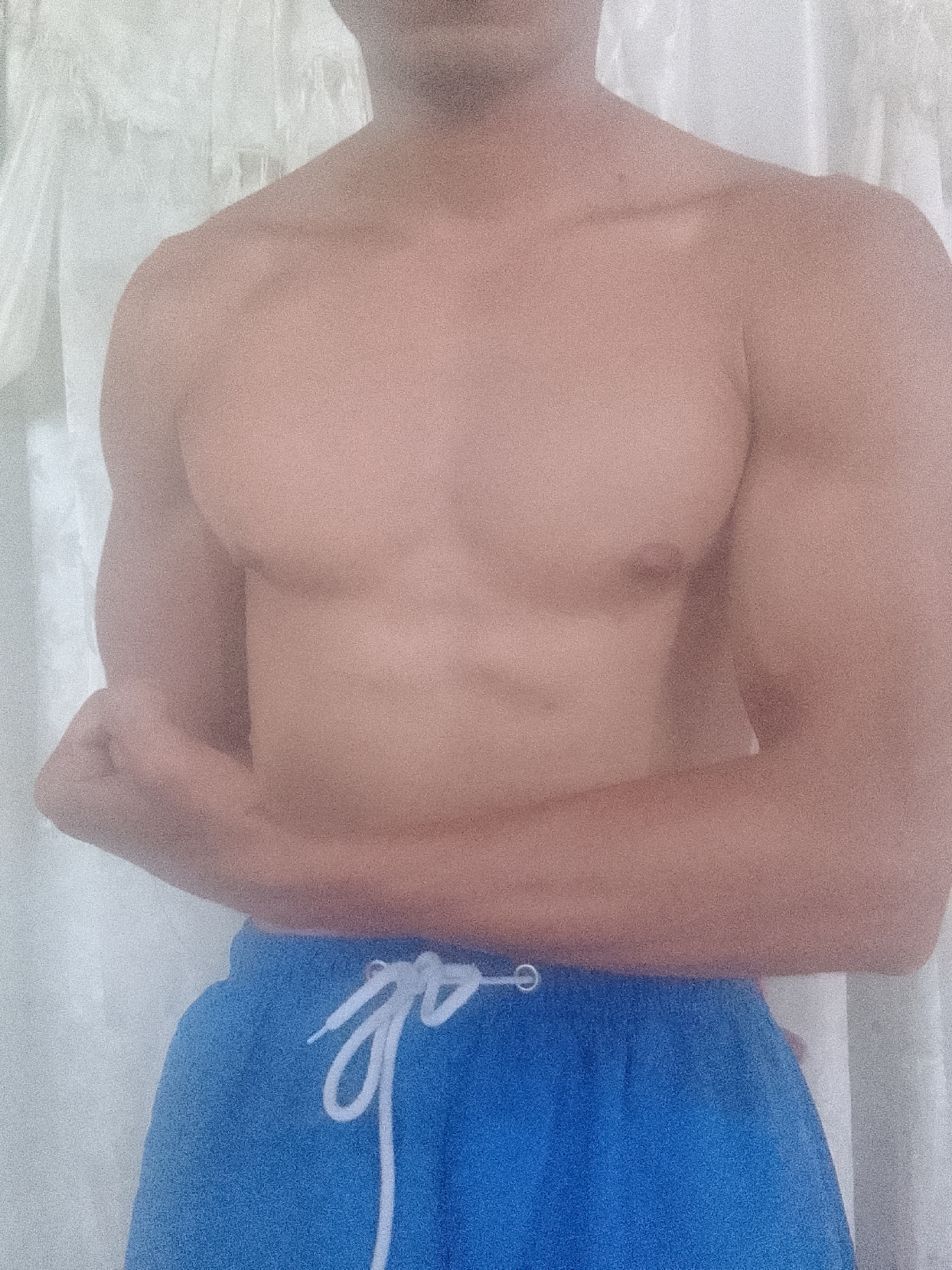 A tanned, shirtless male stands facing the camera in front of a white, textured background. He is wearing blue shorts with white drawstrings. He is flexing his left arm. The location is not discernible from the image, and there is no information to suggest the image was taken in Juan Griego, Venezuela.
