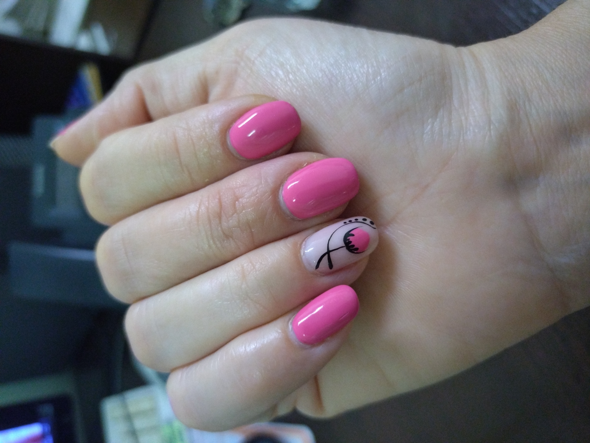 A close-up shot depicts a human hand with manicured nails. The nails are predominantly painted in a bright pink color, except for the ring finger, which features a floral design in black and pink on a light background. The hand is held in a slightly curled position, showcasing the nails. The background is blurred and indistinct, but appears to be an indoor setting. There is no indication of a specific location in Stara Zagora, Bulgaria.