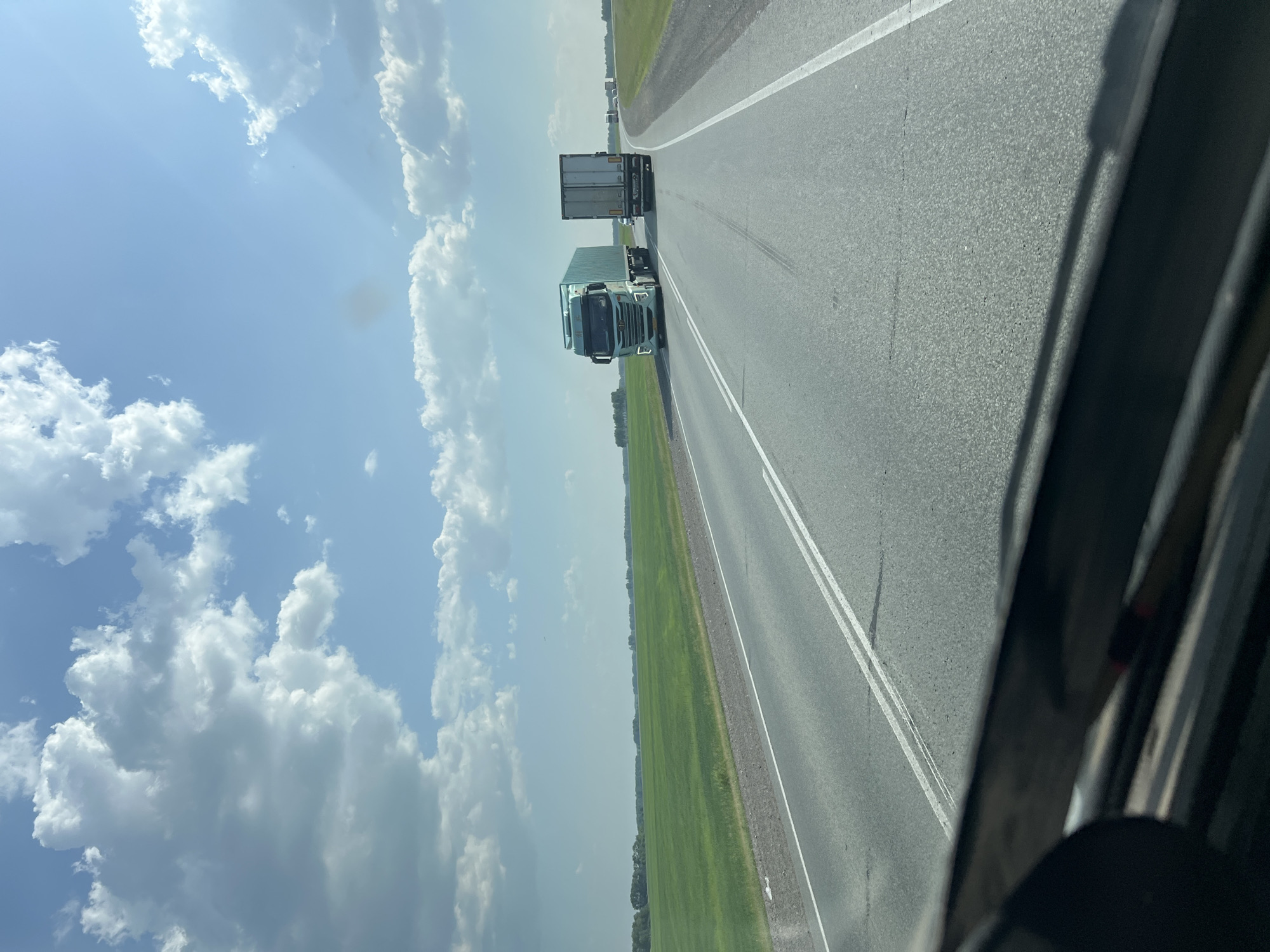 The image is a photograph taken from the driver's perspective of a vehicle traveling on a highway. The highway is paved with asphalt and has white lane markings. To the left of the highway is a vast, green field of crops. In the distance, a line of trees can be seen along the horizon. The sky above is blue with scattered white cumulus clouds.
Two large trucks are visible on the highway. The truck in the foreground is a light blue semi-trailer truck with a white cargo container. The truck behind it is a white semi-trailer truck with a gray cargo container. Several other vehicles, appearing to be cars and trucks, are visible further down the road in the distance, suggesting traffic.
The image appears to be taken in a rural or semi-rural area, given the expansive fields and open sky. The presence of a highway indicates a location with transportation infrastructure. The specific location of Chik, Russia, cannot be determined from the image alone, as the visual elements are generic to many highway landscapes in such regions. There are no people clearly visible in the image.