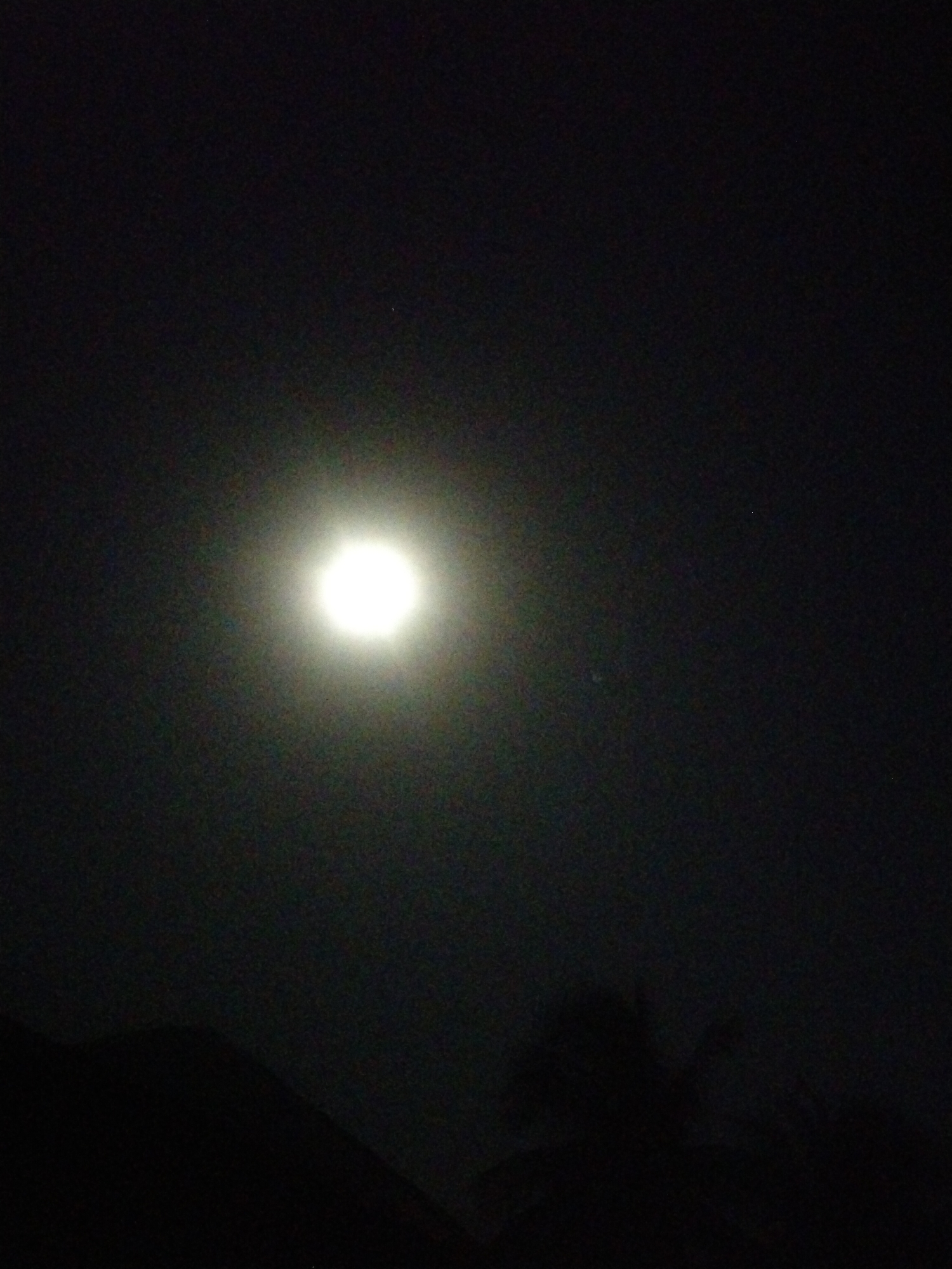 The image shows a night scene with a bright, circular object in the center of the frame, likely the moon. The sky is dark, with some visible gradients suggesting light scattering around the moon. The lower portion of the image shows silhouettes of trees or vegetation, indicating an outdoor setting. There is a darker mass that looks like a hillside in the very bottom left of the image. There are no people visible in the frame. The location is given as Unknown, Unknown.