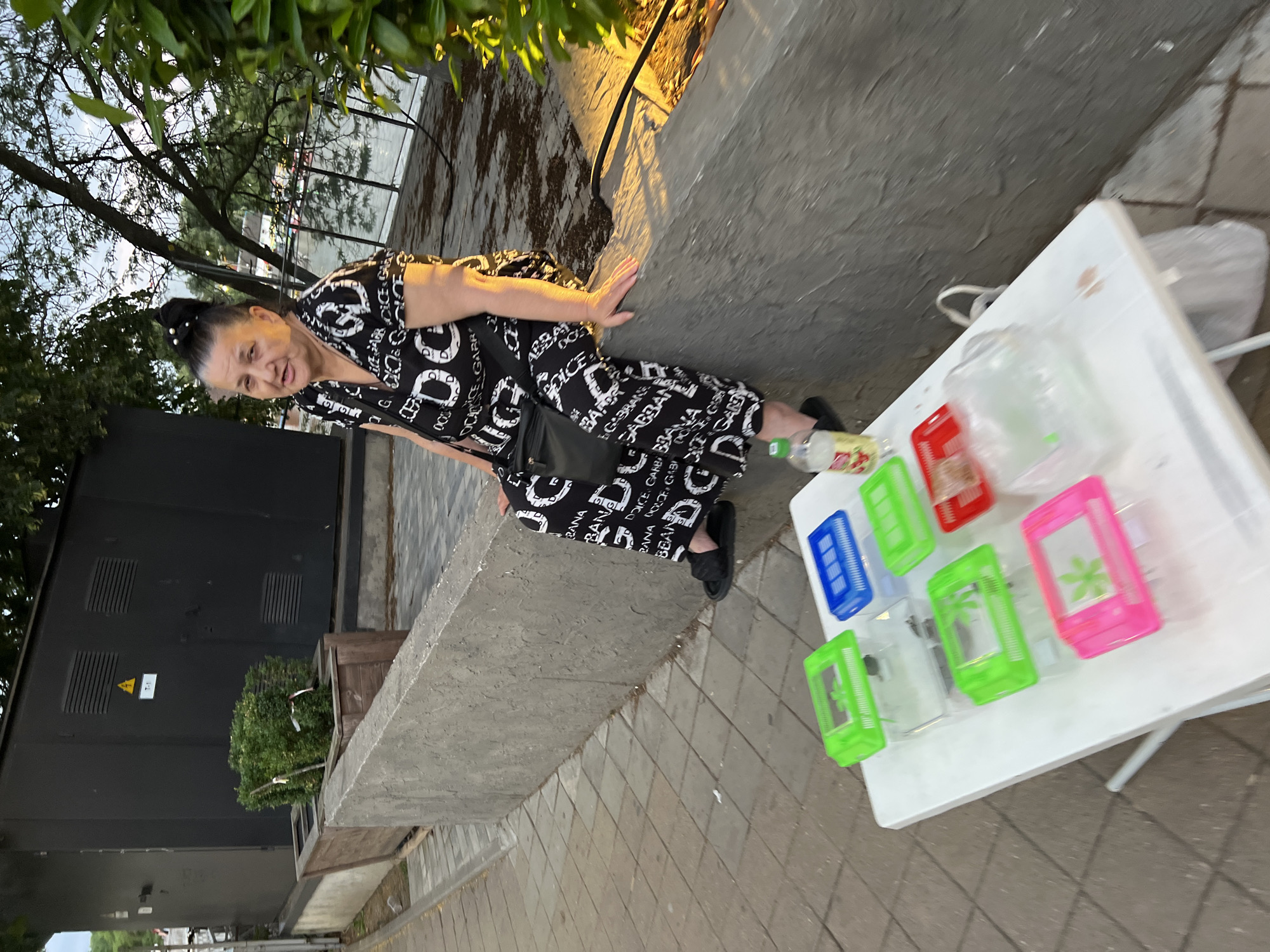 An elderly woman with dark hair styled in a bun is sitting on a concrete barrier. She is wearing a black and white patterned dress and black sandals. In front of her, on a white table, are several clear plastic containers with colored lids, some containing small items. A bottle of liquid and a bag are also on the table. The background shows a body of water with a fence, trees, and buildings. The overall scene suggests an outdoor market or a street vendor setup. The exact location in Krasnodar, Russia cannot be determined from the image alone.