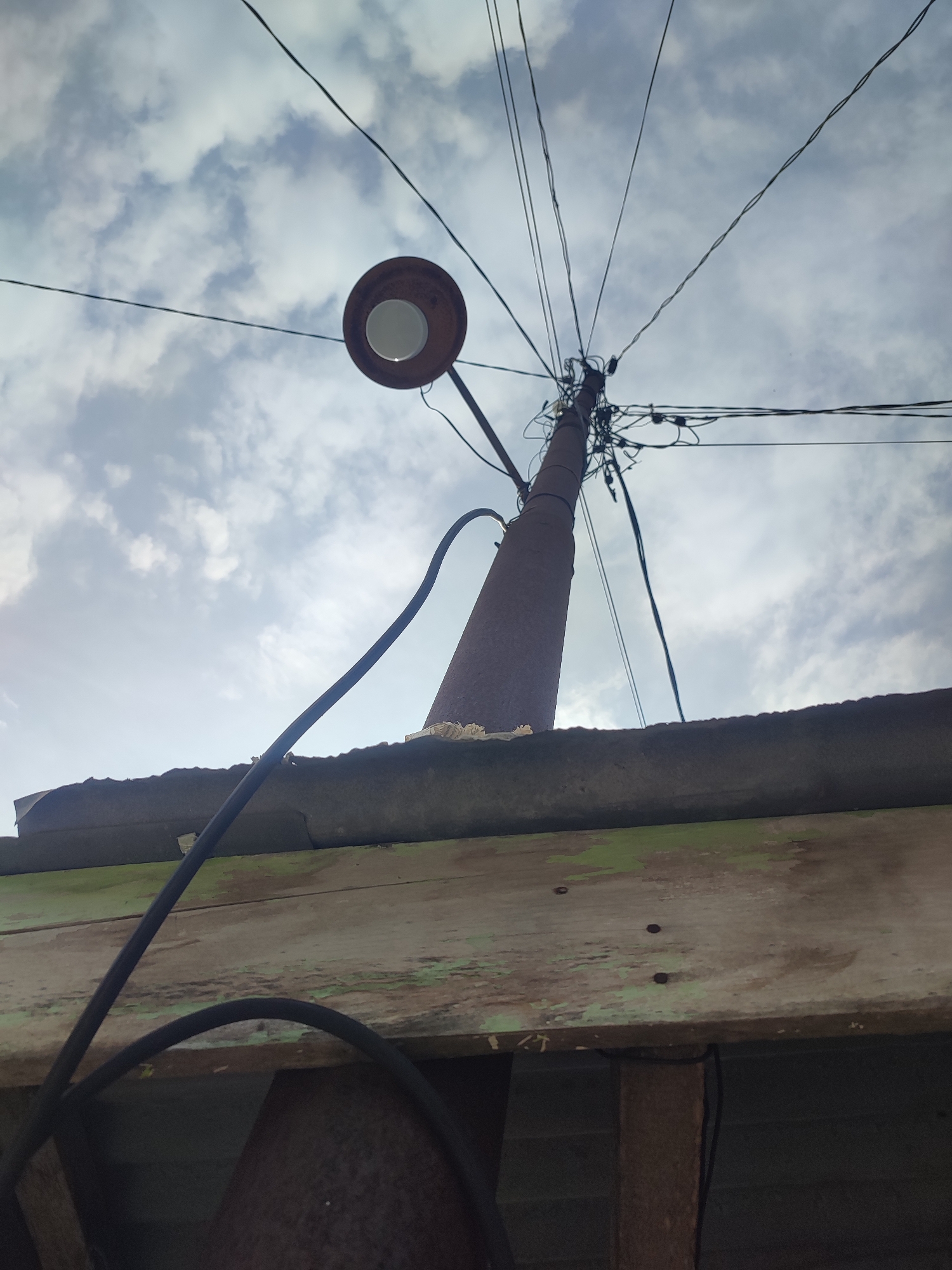 A low-angle shot shows a rusty, conical utility pole in Gampongjawa, Indonesia, supporting a round, overhead street lamp and a chaotic web of power lines against a cloudy sky. The pole emerges from behind the edge of a weathered wooden structure with peeling green paint and a rusty corrugated roof. A thick black cable arcs down from the street lamp, partially obscuring the lower portion of the image. The immediate foreground is dark, obscuring the objects it contains.