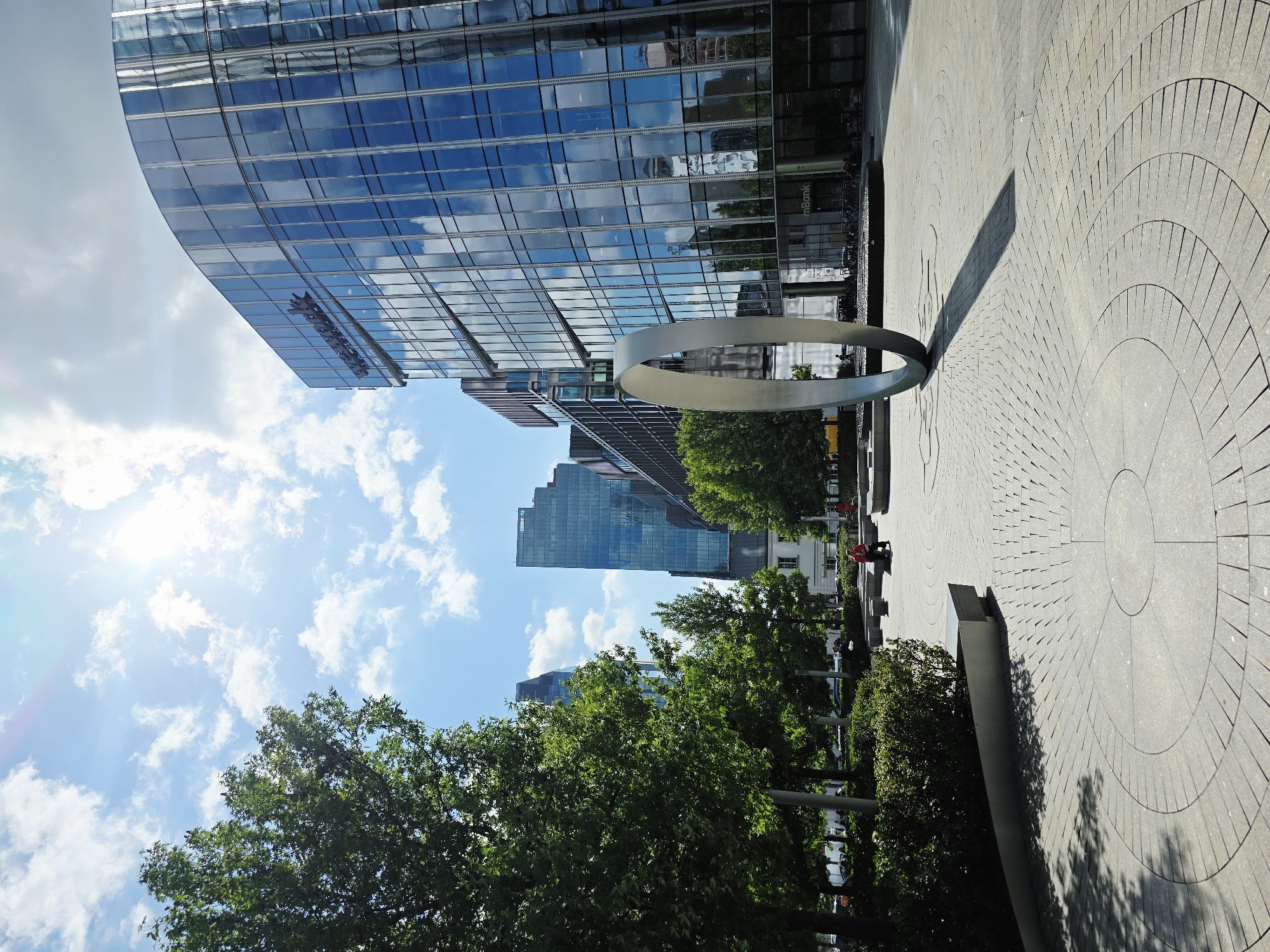 The image shows a modern urban plaza in Warsaw, Poland, likely in the city's business district. The foreground features a circular paving pattern with concentric rings made of gray stone. A large, metallic, ring-shaped sculpture stands prominently in the plaza, casting a long shadow. To the right, a multi-story building with a curved glass facade reflects the sky and surrounding architecture. The "mBank" logo is visible on the lower portion of the building. In the background, several tall office buildings with glass exteriors rise into a bright blue sky filled with scattered white clouds. Lush green trees are planted around the plaza, and a few people are visible sitting on benches and walking. The overall impression is one of a clean, contemporary, and well-maintained public space in a bustling city.