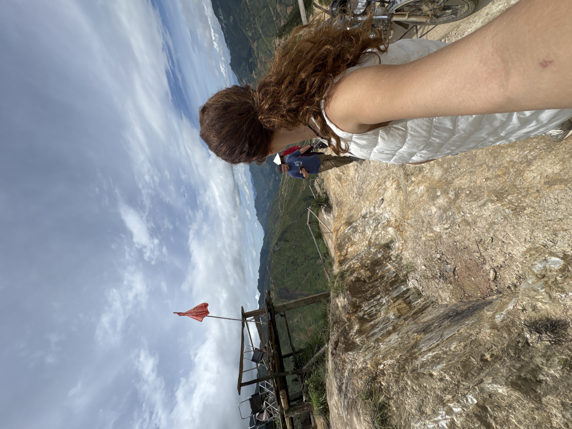 A woman with long, curly auburn hair tied in a ponytail, wearing a white puffer vest, takes a selfie from a selfie angle as she stands on a rocky, uneven path. Behind her, the path leads towards a wooden platform with chairs. A red flag is planted on the platform, which overlooks a vast, green, mountainous landscape under a cloudy sky. Two other individuals, one in a blue shirt and another in a red shirt, are visible further down the path. A bicycle is parked to the right of the woman. The scene appears to be in Bắc Yên, Vietnam.
