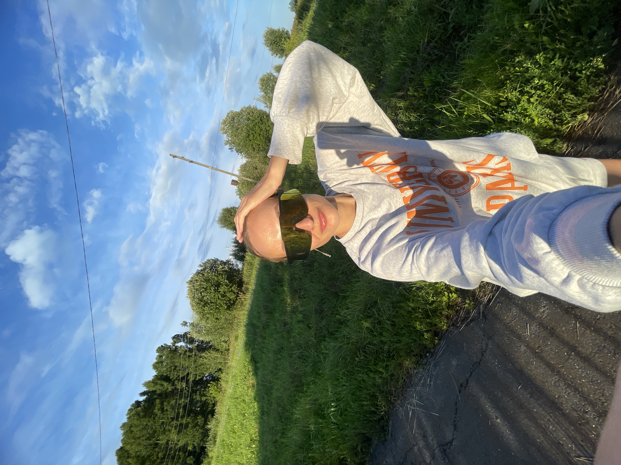 In the image, a woman is taking a selfie outdoors in a rural setting, likely in or near the village of Podshivalovo, Russia. She is wearing a grey sweatshirt with orange lettering that appears to say "UNIVERSITY DAYS" and large, tinted sunglasses. Her right hand is resting on the top of her head. The background consists of a grassy hill with some trees and vegetation, and a bright blue sky with scattered clouds. A power line is visible stretched across the sky. The lighting suggests it is late afternoon or early evening, with the sun casting shadows. The ground in the foreground is a dark, cracked path, possibly asphalt.
