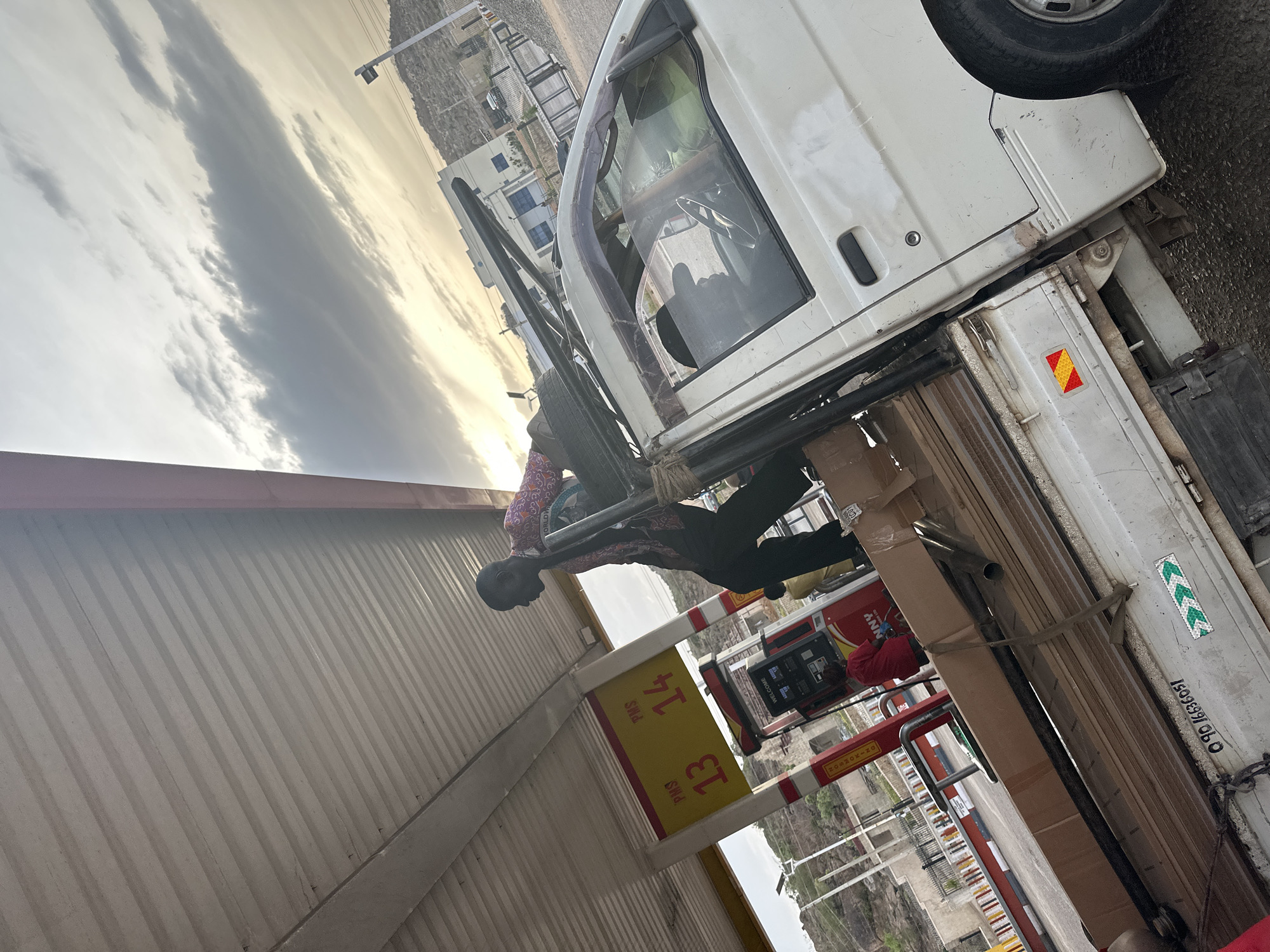 The image, taken in Gura Top, Nigeria, depicts a scene at what appears to be a gas station or service area under an overcast sky, possibly during late afternoon or early evening as suggested by the soft light and golden hue on the horizon. The primary subjects are a white pickup truck or commercial van and a man working near its rear.
The man, dressed in a patterned long-sleeve shirt and dark trousers, is positioned on the driver's side of the truck, appearing to be engaged in loading, unloading, or securing cargo. The truck's bed is laden with several large, rectangular cardboard boxes and what look like pipes or rods, secured with straps. The truck itself shows signs of wear, with visible dirt and minor damage on its white body. A spare tire is mounted on the side.
To the left of the man and truck, a yellow sign with red lettering is visible, displaying "PNS 13" and "PNS 14," likely indicating pump numbers or service points. Behind this, a red gas pump with the text "ANN" and "WELCOME" is partially visible, with another person in a red shirt standing nearby, facing away from the camera. The gas station features a white, corrugated metal canopy overhead.
In the background, a rugged, mountainous or hilly landscape stretches out under the cloudy sky. Some simple buildings are visible in the distance, nestled against the hills. Notable details on the truck include a red and yellow reflective hazard sticker and a green and white chevron pattern. A number "090 16686051" is also printed on the side of the truck's cargo bed. The overall scene suggests everyday activity at a roadside stop in a rural or semi-urban setting.