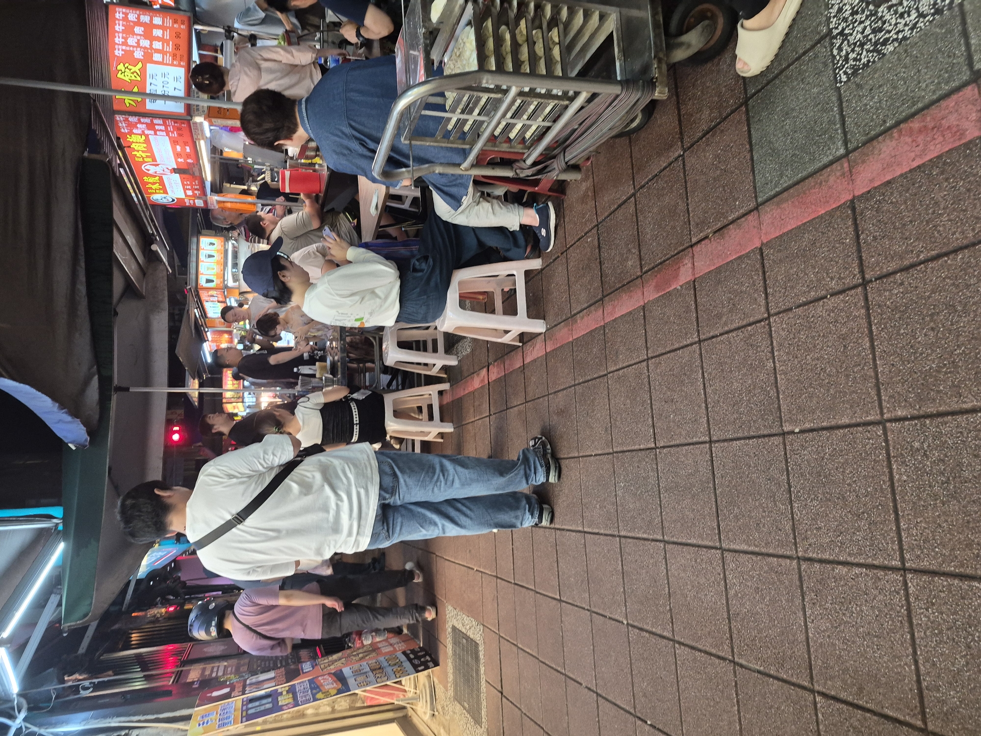 This is a medium-shot, eye-level outdoor photograph taken at night on a tiled street in Taipei, Taiwan. The scene depicts a busy street food stall area with several people engaged in various activities.
In the foreground, a person wearing a white t-shirt and blue jeans stands with their back to the camera, facing away from the viewer. They have a dark strap across their chest. To their left, a person in a purple t-shirt and dark pants walks away from the camera, wearing a helmet.
Several individuals are seated at tables in the midground. One person in a white long-sleeved shirt and dark pants is looking down at their phone. Another person in a denim jacket is seated at a table. A person in a black and white top is sitting on a white stool. Further back, more people are visible at tables and standing.
The background features several food stall signs, some of which are illuminated with red and white lettering, indicating menu items and prices. A metal cart with multiple shelves stacked with food items is visible on the right side of the frame. The ground is paved with reddish-brown square tiles, with a thin red line marking a boundary on the pavement. The overall impression is of a lively, commercial street environment at night.