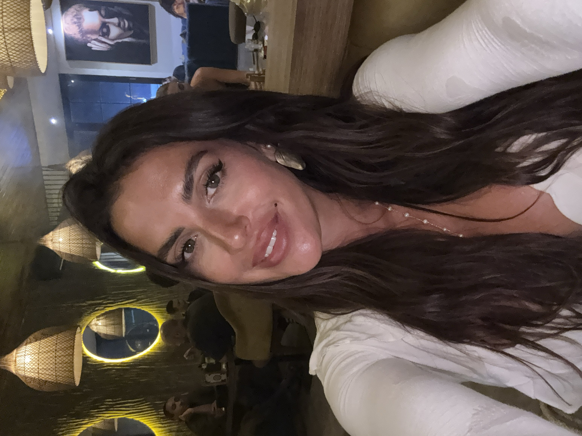 An indoor, selfie-style photograph shows a woman in the foreground. She has long, dark, wavy hair and is wearing a white top with a textured, layered neckline and sleeves. She has on a delicate necklace with pearls. She is wearing large, leaf-shaped earrings. Her eyes are open and she is looking at the camera, with her lips parted in a smile, revealing her teeth. Her makeup includes eyeliner and lipstick. She has a mole below her lower lip.
The background suggests an indoor dining or lounge area, possibly a restaurant. There are several people seated at tables in the mid-ground. The lighting is dim and warm, with textured lampshades hanging from the ceiling. There are round, illuminated mirrors on the wall. On the far right, a framed artwork depicting a woman's face is visible. The overall ambiance is dimly lit and sophisticated. This scene is located in Kuta Selatan, Indonesia.