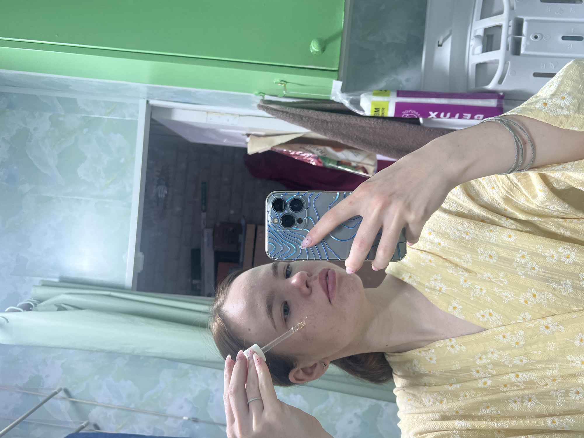 A young woman is taking a selfie in what appears to be a bathroom. She is holding a phone with a blue patterned case in her right hand and a dropper in her left, applying a liquid to her face. She is wearing a yellow dress with a white floral pattern and several silver bracelets. The bathroom has green cabinets and patterned wallpaper. Behind the woman, there is a shelf with various items, including a purple box with Cyrillic text. The overall impression is of a personal grooming routine being documented. There is no information to indicate this image was taken in the city of Chaikovsky, Russia.