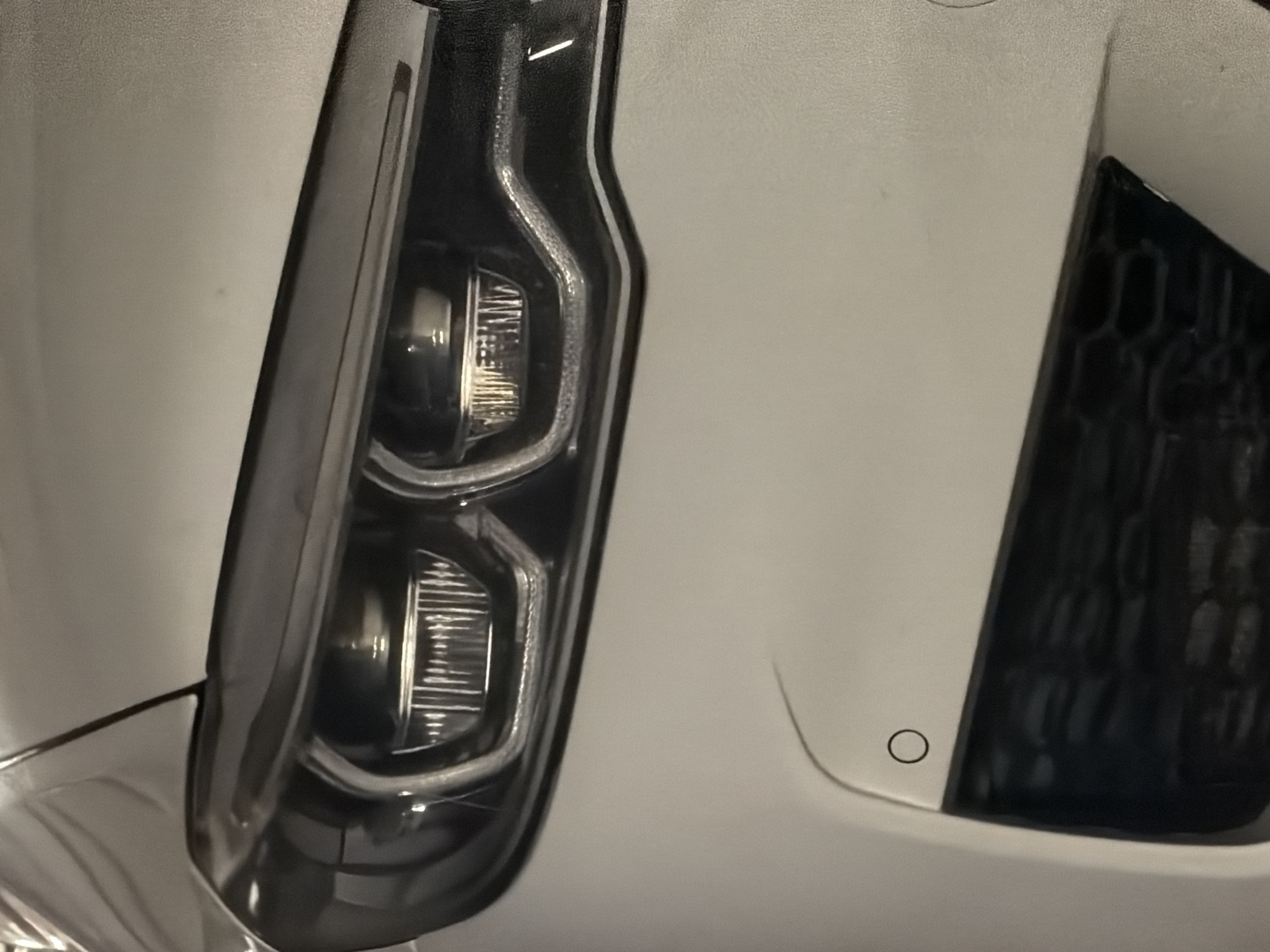 This image depicts the front right section of a white passenger vehicle, located in Burgas, Bulgaria. The primary subject is the vehicle's right headlight assembly, which is illuminated. The headlight unit features two distinct projector lenses: one circular and one with a faceted rectangular lens, both actively emitting light. A bright, curvilinear LED daytime running light strip outlines the lower and outer perimeter of the headlight housing. Below the headlight, a white bumper is visible, integrating a single circular parking sensor positioned on its surface. A section of a dark, hexagonal mesh grille or lower air intake is present beneath the bumper line. The upper portion of the vehicle's white hood and fender are also visible. The scene is static, devoid of any individuals, actions, or interactions. The lighting is uniform and artificial, indicating an indoor environment such as a parking garage or showroom.