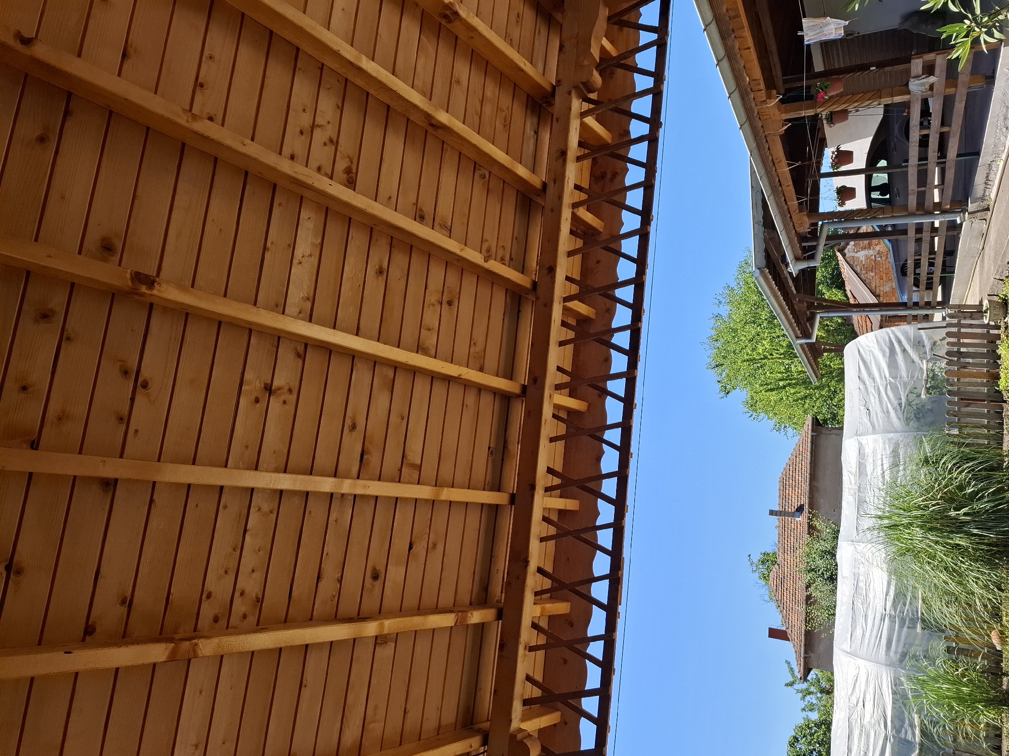 The image shows an outdoor scene beneath a wooden-slatted roof with exposed wooden beams. The roof is supported by wooden posts. The sky is clear blue. In the background, there is a house with a red-tiled roof and two chimneys. To the left of the house, there is a structure covered with white plastic sheeting, possibly a greenhouse. To the right of the house, there are two adjacent wooden structures with roofs and fences. A grey car is parked under one of these structures, and hanging flower pots are visible. This scene is located in the city of Galabovo, Bulgaria.