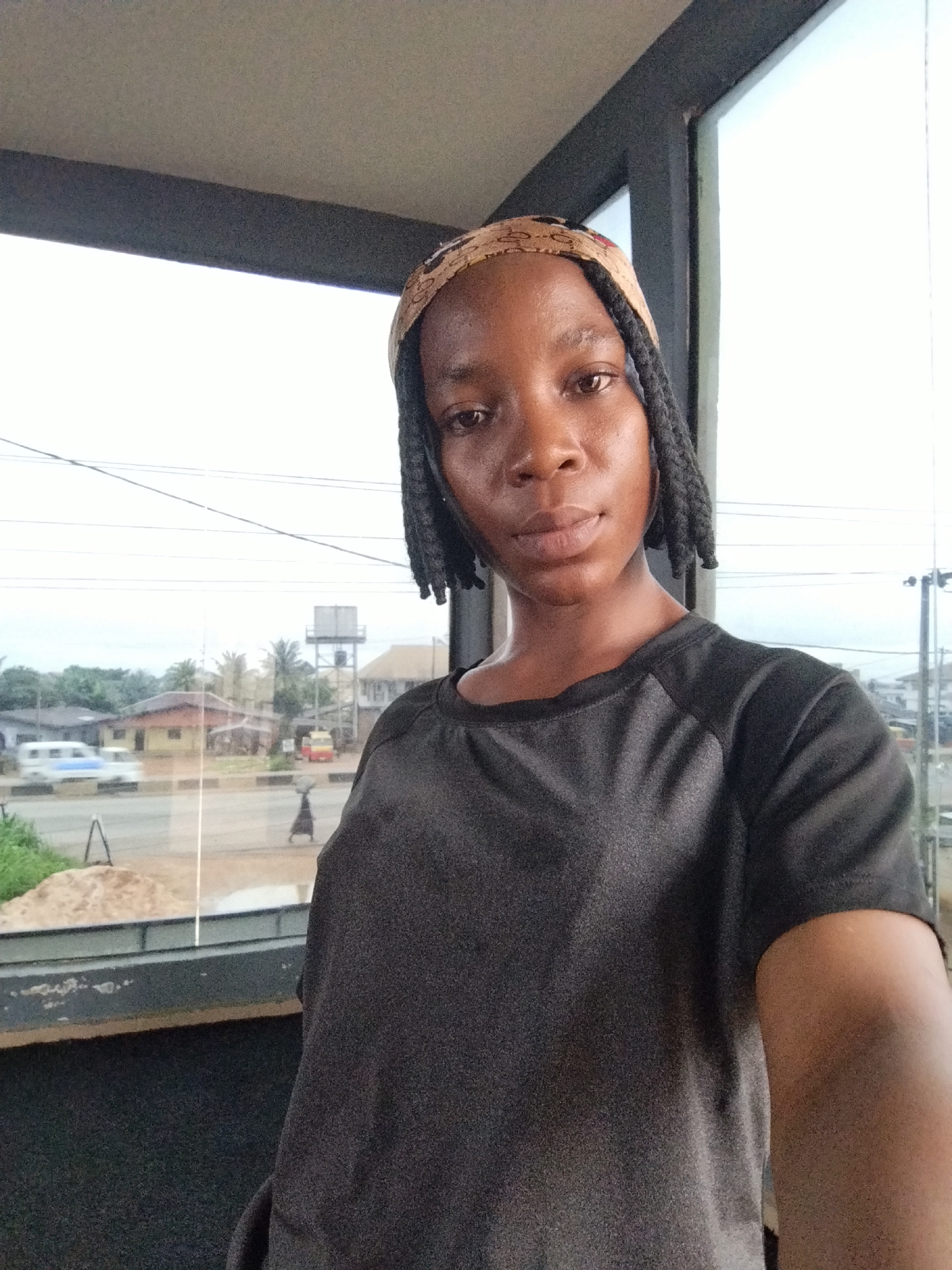 A young Black woman with dark skin and braided hair, wearing a dark grey t-shirt and a patterned headband, is positioned in the foreground of the image. She is looking directly at the camera. Behind her, a large window reveals a street scene in Uselu, Nigeria. Several buildings, including houses and a water tower, are visible, along with trees and a road with some vehicles, including a white van and a smaller red vehicle, suggesting a typical daytime environment in the area. The lighting indicates it is daytime.
