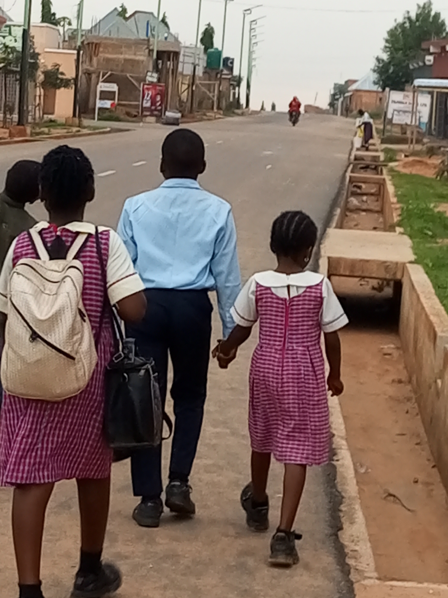 A medium shot captures three children and one adult walking away from the camera on a paved road in Tempure, Nigeria. The children, two girls and a boy, are dressed in school uniforms. The older girl on the left wears a pink and white checkered dress over a white shirt and carries a beige backpack. The boy, in the middle, wears a light blue collared shirt and dark trousers. The younger girl on the right wears a similar pink and white checkered dress over a white shirt. All three children are walking on the left side of the road, away from the camera. The adult, a man, walks to the right of the boy, also facing away from the camera, wearing a light blue collared shirt and dark trousers.
The road appears to be a two-lane street with white dashed lines marking the lanes. The area to the right of the road features a drainage ditch and a grassy embankment with scattered vegetation. In the background, buildings with corrugated metal roofs and some shops are visible. A motorcycle carrying a rider in a red garment is moving away from the camera in the distance on the road. The sky is overcast, indicating diffused lighting conditions. The scene suggests a typical urban or semi-urban environment in Nigeria, possibly during a school day.