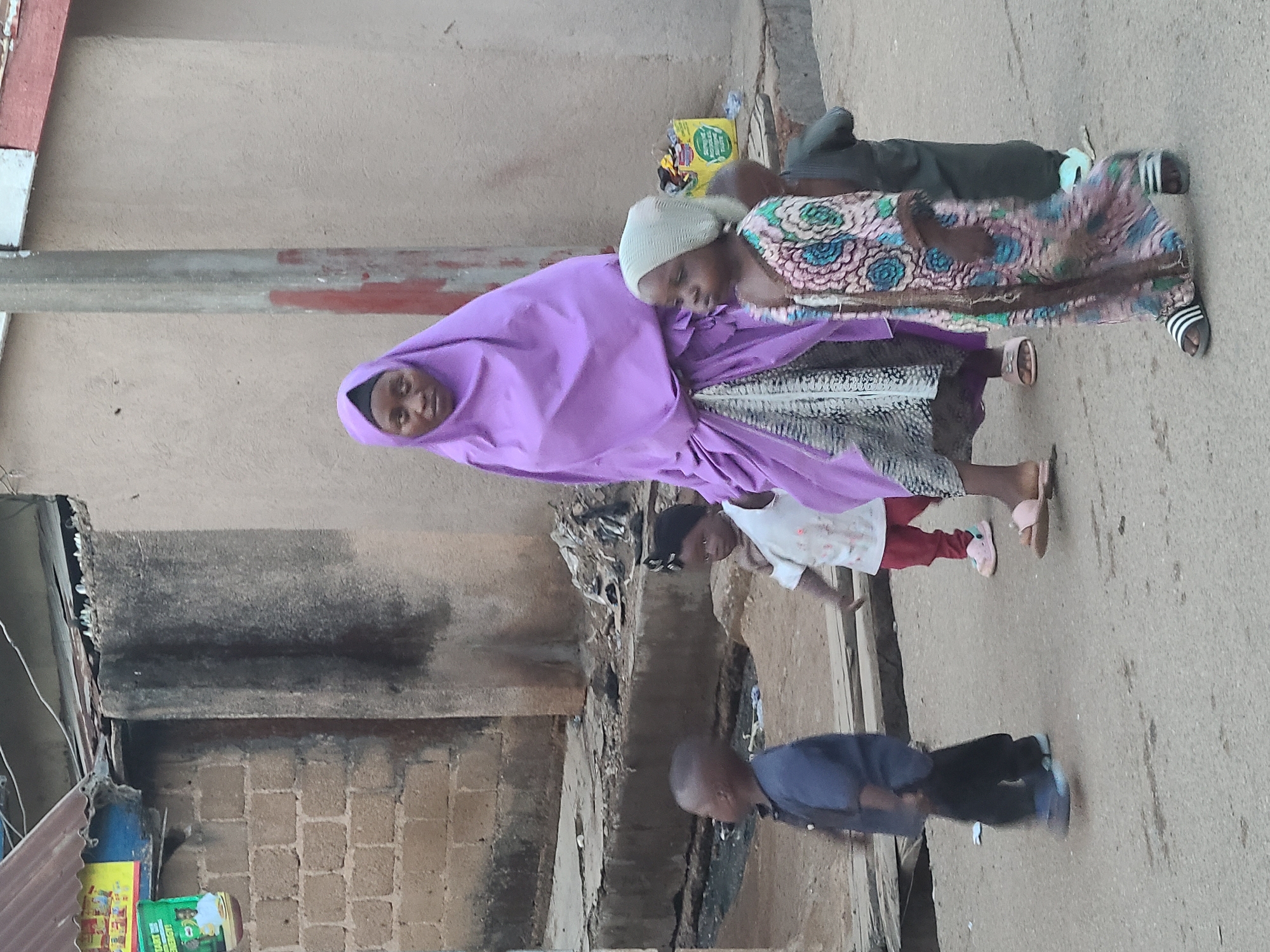 This image captures a candid moment of five individuals outdoors, likely on a street or alleyway. In the foreground, a woman or older girl stands centrally, wearing a light purple hijab that covers her head and shoulders, and a patterned skirt or dress. She looks calmly towards the viewer. Next to her, a young girl with a light head covering and a colorful patterned dress leans her head against the older woman's shoulder, appearing to rest or be comforted.
Behind this pair, another child in a dark top and trousers is partially visible, wearing striped sandals. Further to the left and slightly behind, a child in a white top, reddish trousers, and pink sandals is visible. In the immediate foreground, a very young child or toddler, dressed in a dark blue top, dark trousers, and blue shoes, stands on the paved ground, looking downwards.
The setting appears to be a residential area with buildings made of brick and plastered walls, some showing signs of wear and tear. The ground is paved or concrete, and a narrow gutter or drain runs across the mid-ground. On the far left, corrugated metal sheets are visible. A yellow container with some unreadable green text, possibly "START" and "ENERGY", is present on the left side of the scene, along with other debris. The image is taken during the daytime under soft or ambient lighting. The clothing, particularly the hijabs and sandals, suggests a location in a warm climate, possibly within a community where Islamic attire is common.