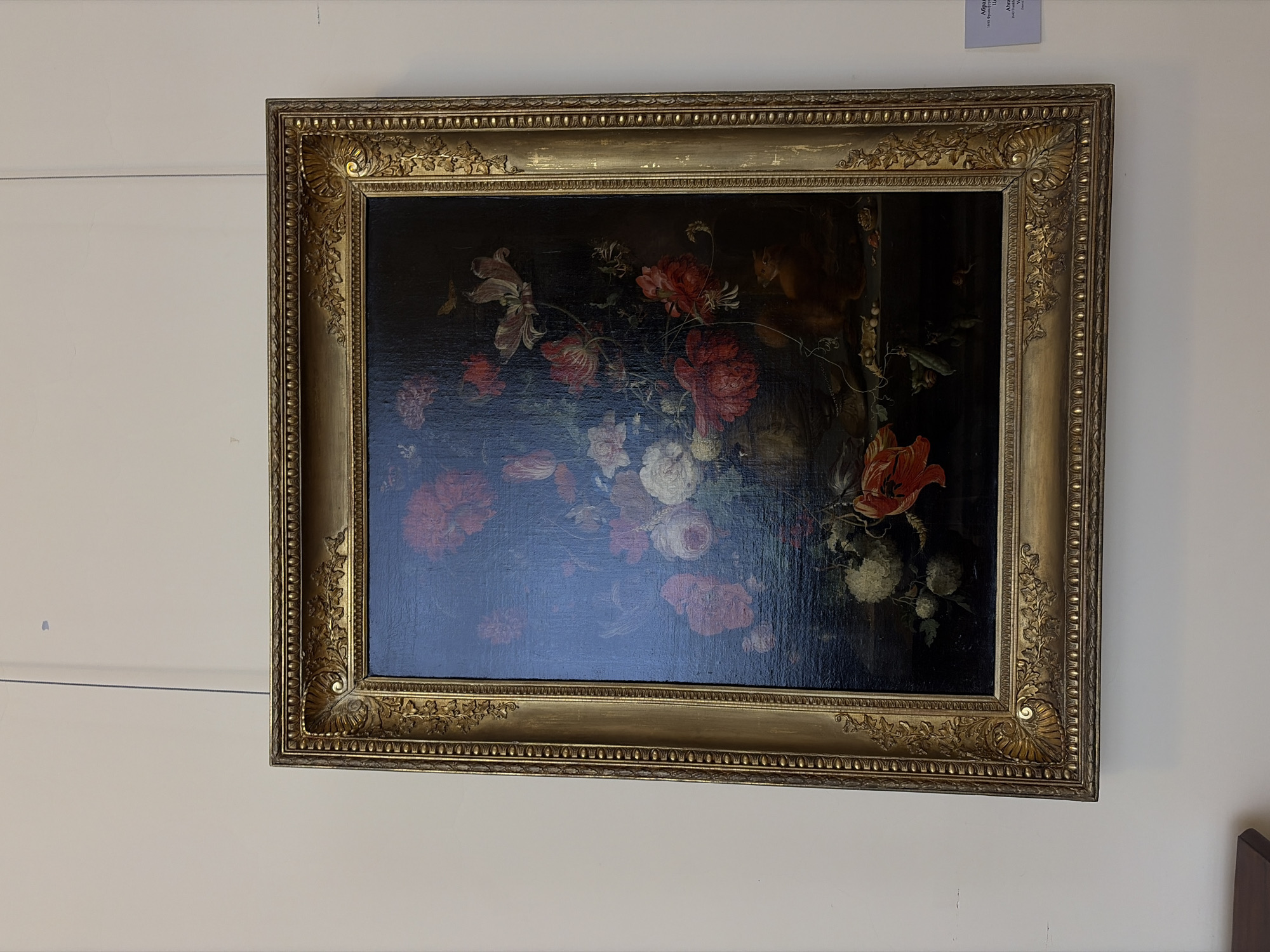 A still-life painting of flowers hangs on a white wall. The painting is framed in a wide, ornate gold frame. The painting depicts a dark background with a colorful arrangement of flowers in shades of red, pink, and white. The scene appears to be indoors, with the painting hung at eye level. The location is Saint Petersburg, Russia.