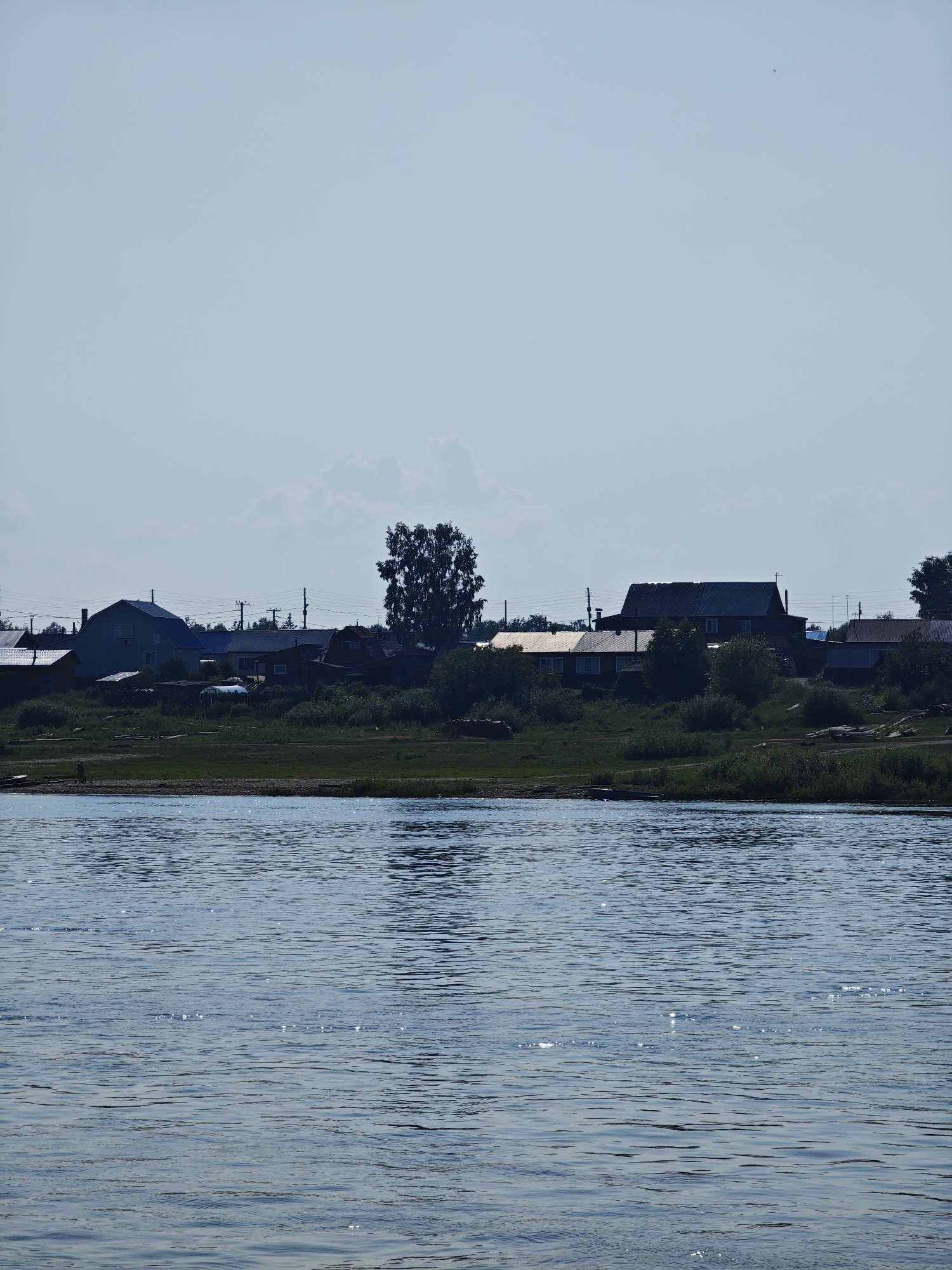 A wide, calm river with a slight ripple effect occupies the foreground of the image. The water is a muted blue-gray and reflects the light of the sky, creating shimmering highlights. In the middle ground, a densely wooded riverbank is visible, transitioning to a small village nestled on a slightly elevated area. The village consists of several houses with pitched roofs, some appearing to be wooden structures, while others are painted in lighter colors. Power lines and poles run along the edge of the village. A large, dark silhouette of a tree stands prominently to the right of the village. The background is a hazy, light blue sky with a few soft clouds. The overall impression is of a serene, rural landscape.
