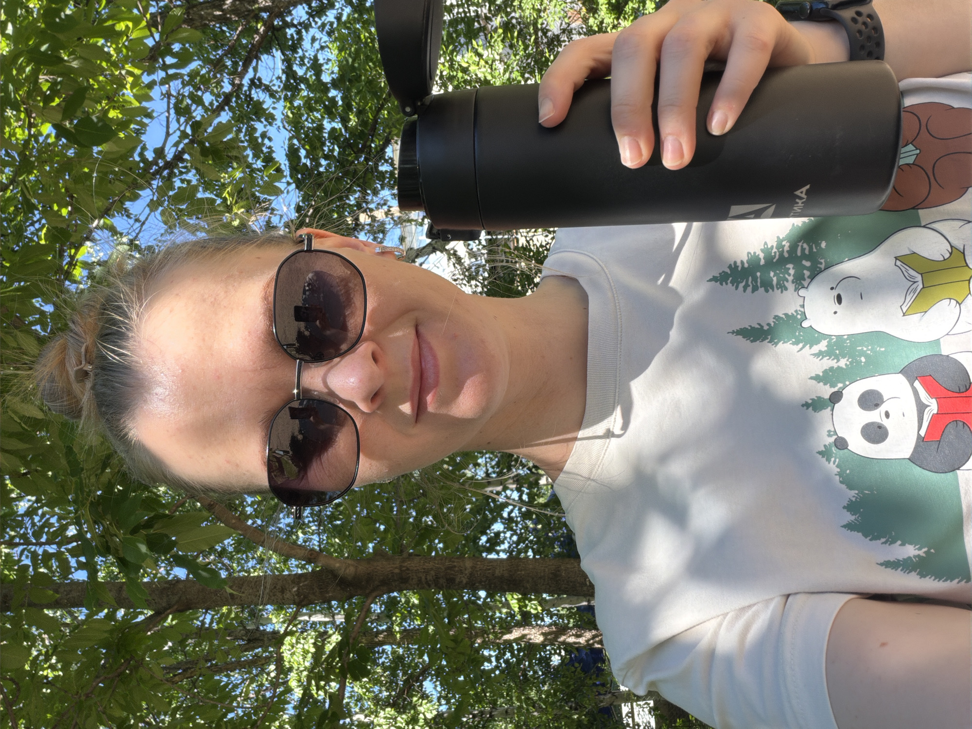 A young Caucasian woman with blonde hair pulled into a bun wears a light-colored t-shirt with a graphic of three cartoon bears and pine trees. She is holding a black insulated water bottle in her right hand and is wearing dark-rimmed sunglasses. The background consists of green foliage and a blue sky, suggesting an outdoor setting. The image was taken on a sunny day in Khabarovsk, Russia.