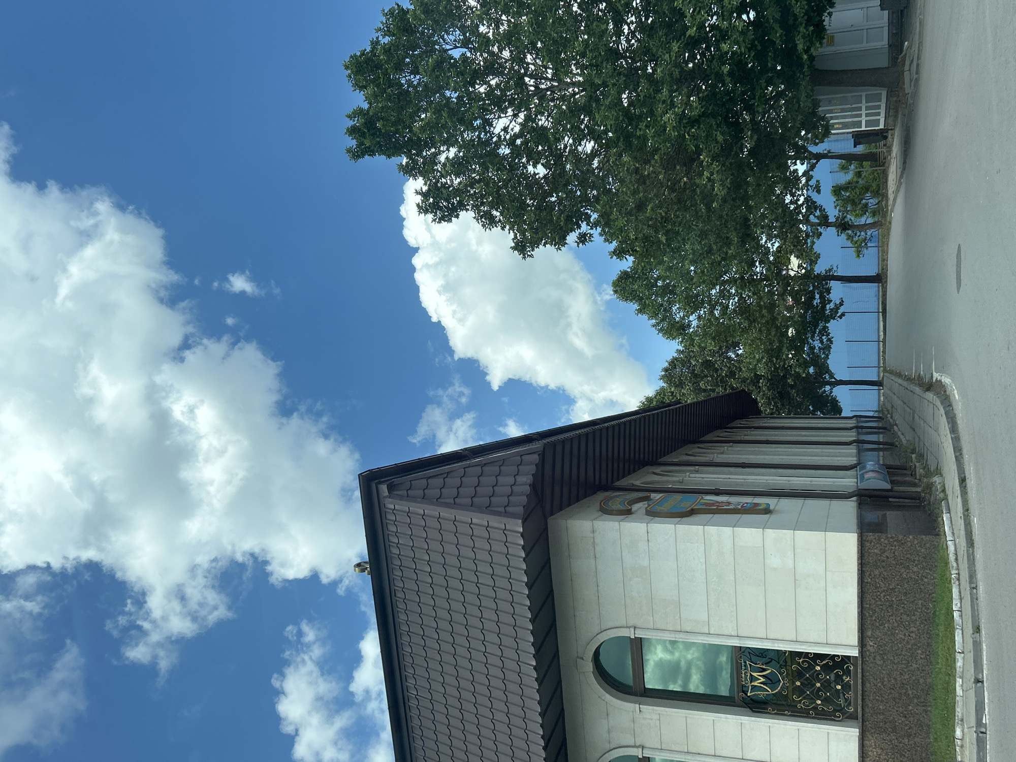 This detailed image captures a building and street scene in Sofia, Bulgaria, specifically identifying the structure as **Hotel Rotasar**, located adjacent to Arena Sofia (formerly Mtel Arena) in the Izgrev district.
The scene unfolds on a bright, sunny day under a vivid blue sky dotted with numerous white, cumulus clouds. In the foreground and dominating the left side of the frame is a multi-story building with a distinctive architectural style. Its facade is constructed from light-colored, possibly beige or cream-toned, large stone blocks, featuring a darker, speckled brown granite base. The building boasts a steep, multi-faceted roof covered in brown, interlocking tiles, creating a textured surface. A small, metallic, curved decorative finial or element is visible at the apex of the roofline.
Several arched windows with dark frames are visible, prominently reflecting the blue sky and white clouds, giving them a mirrored appearance. One such window on the front-left corner of the building features an elaborate black wrought-iron grate with intricate scrollwork. At the center of this grate, a golden crest or emblem is displayed, featuring a stylized capital "M" within a decorative shield. Along the right side of the building, a series of vertical architectural pillars or pilasters extend into the background, and beneath them, some indistinct, rectangular signs or posters are visible at ground level.
An asphalt road occupies the lower portion of the image, curving gently to the right as it recedes into the distance. A paved sidewalk or pedestrian path runs alongside the building and the road. To the right of the road, a line of mature deciduous trees with lush green foliage stands tall, casting dappled shadows. Beyond these trees, a prominent, solid light blue barrier or fence stretches horizontally across the background, suggesting the boundary of a larger property or complex. Further to the far right, a small, white, utilitarian structure with a door and window is partially visible. No people or active interactions are present in the frame.