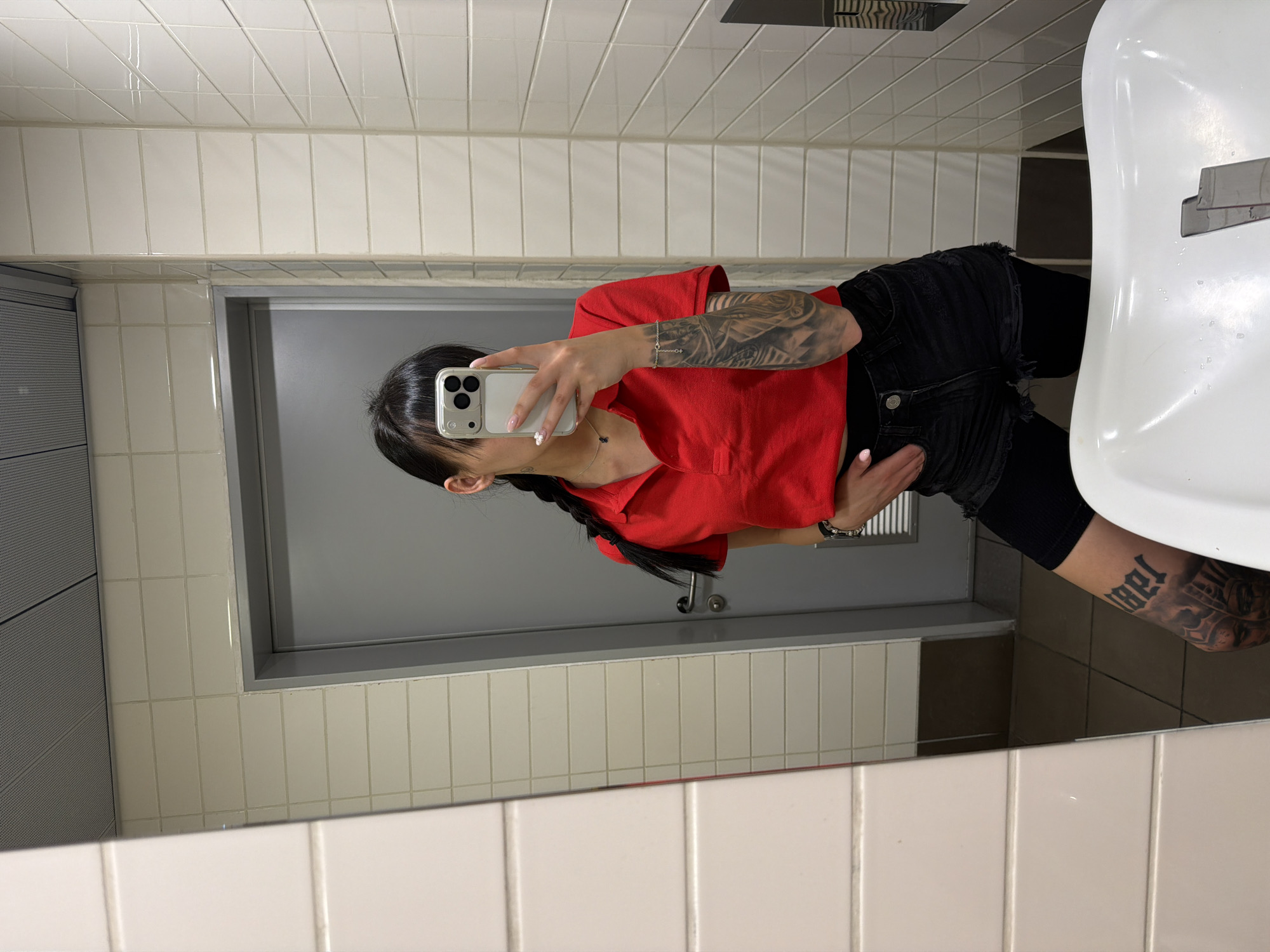 A young woman with dark hair in a braid is taking a mirror selfie in a public restroom. She is wearing a red polo shirt, black denim shorts, and black leggings. Her left arm is heavily tattooed, and she has tattoos on her right thigh. The restroom has white tiled walls and a gray door. A white sink is visible in the foreground. The lighting is bright and even.