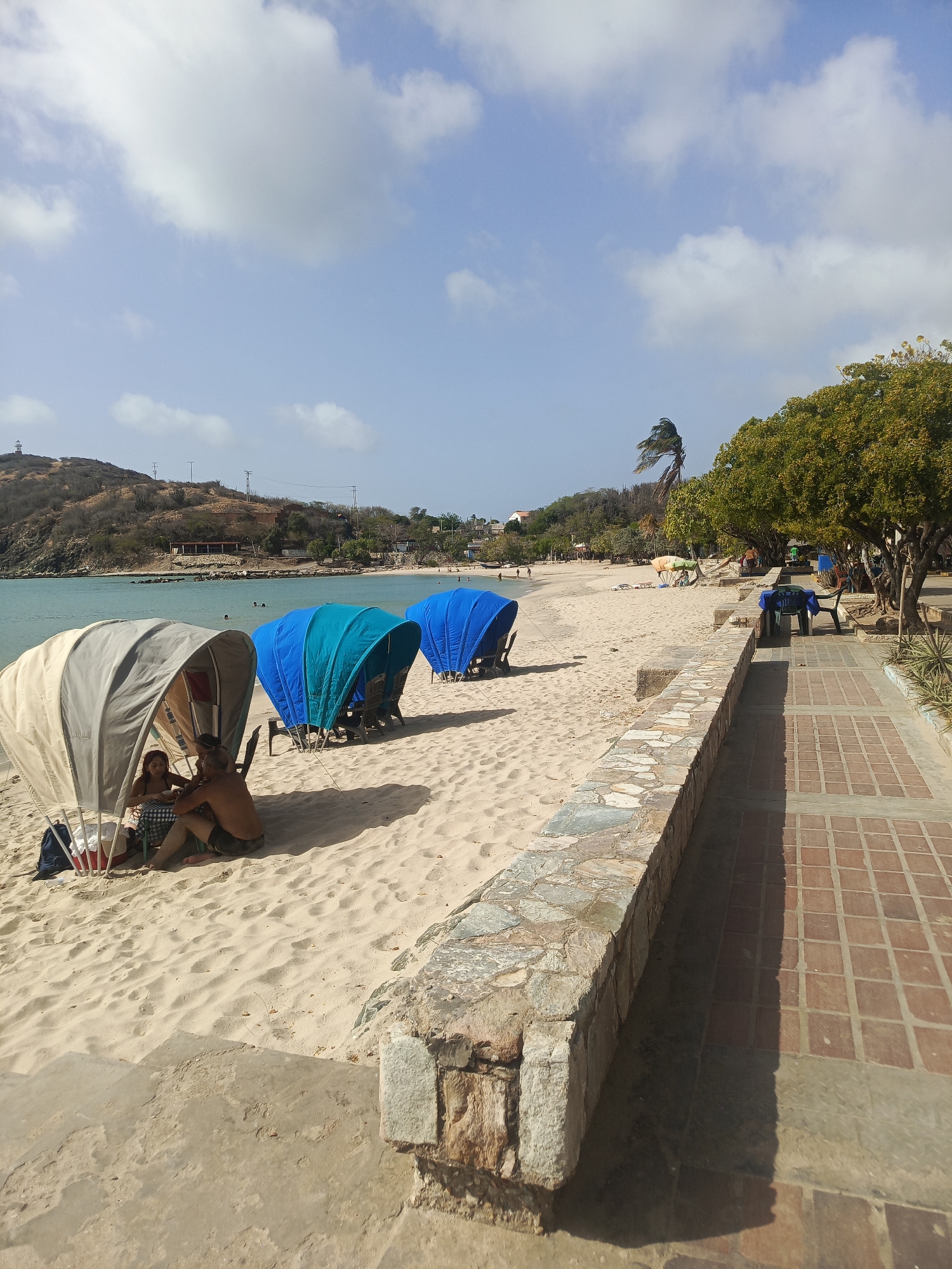 This is an outdoor scene at a beach in Pedro González, Venezuela on a sunny day with scattered clouds. The beach is light sand, and calm teal water is visible in the background.
In the foreground, there are three covered seating areas with chairs. The first is tan, and two people are sitting inside. The next two are teal and blue.
A low stone wall separates the beach from a brick walkway along the right side of the image. On the walkway, there are tables and chairs beneath trees. In the distance, the landscape consists of hills and buildings.