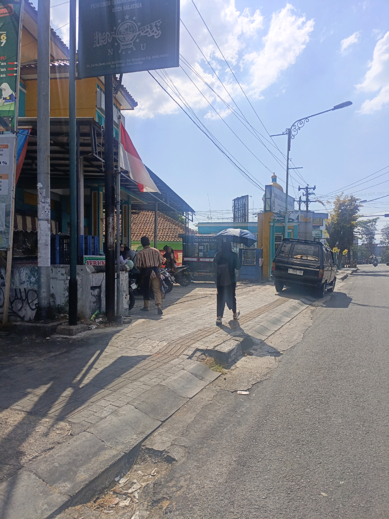 The image depicts a street scene in the city of Salatiga, Indonesia, under a bright, partly cloudy sky. In the foreground, a paved sidewalk runs alongside a road. Several people are visible. On the sidewalk, a person is walking away from the camera, holding a dark umbrella over their head. Another person is walking behind them. Closer to the buildings, two individuals are seated on a motorcycle, with another person standing nearby. A parked black van with a license plate reading "H 1935 GT" is on the right side of the road.
Several buildings are present in the background. On the left, a two-story structure has a yellow facade with an Indonesian flag draped from an upper level. Above the entrance of one building, a sign displays a logo and text that appears to be in Arabic script, followed by "N U" and "KOTA SALATIGA". Another sign, partially obscured, has the words "SAN DI". A street lamp is visible in the mid-ground, and utility lines crisscross the sky. Trees and other foliage are sparsely scattered throughout the scene. The ground on the sidewalk and along the roadside shows signs of wear and some litter. The overall impression is that of a typical urban street on a sunny day in Indonesia.