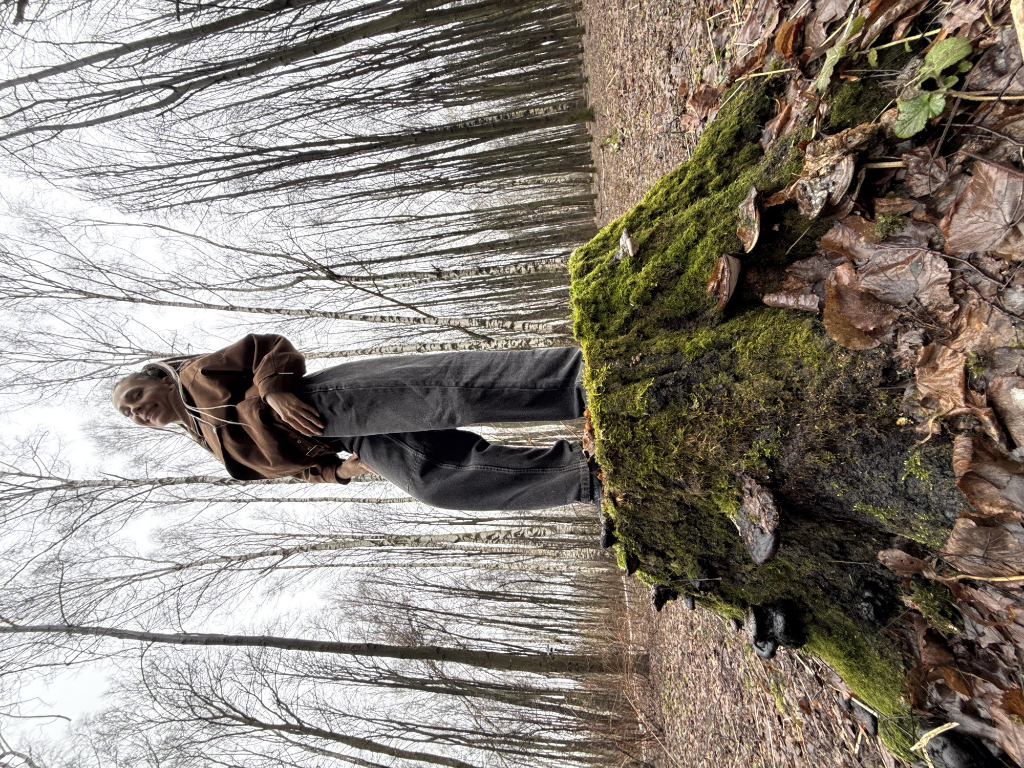 A low-angle, rotated shot shows a person in a brown jacket and black jeans standing on a moss-covered tree stump in a forest. The person, fair-skinned with light hair, is positioned on the left side of the stump, looking towards the camera with a slight smile. The stump is covered in green moss and decaying leaves, and there are what appear to be mushrooms or fungi growing on it. The background is filled with rows of tall, leafless trees, creating a forest setting under an overcast sky. The ground is covered in fallen leaves and twigs. The precise location within the city of Unknown, Unknown is indeterminable.
