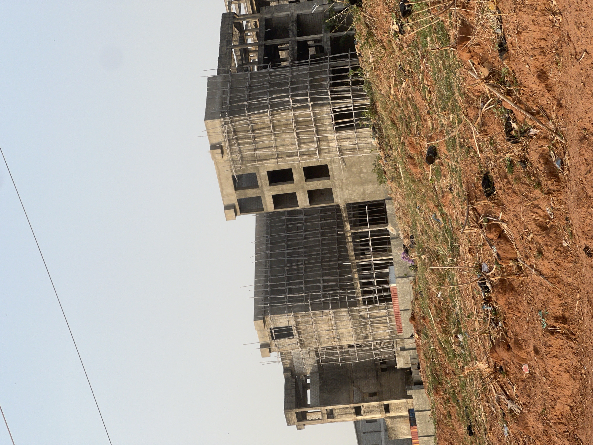 This image depicts a construction site in Abuja, Nigeria, under a bright, clear sky. In the foreground, a reddish-brown earth covers the ground, sparsely covered with dry, light-colored grass, weeds, and scattered debris including dark plastic bags, dried branches, and some litter.
The mid-ground and background are dominated by several multi-story concrete buildings in various stages of construction. The most prominent building on the right features a raw concrete frame with numerous square and rectangular openings that will eventually be windows or balconies. A complex network of scaffolding, made from slender wooden poles, is affixed to the exterior of this building, indicating active or recently paused construction work. Another uncompleted concrete structure is partially visible further to the left, also showing exposed concrete and rebar. The buildings appear to be shells, lacking finishes, windows, or doors.
The sky is a light blue, completely clear and cloudless, suggesting a bright day, likely late morning or early afternoon, with good visibility. A thin, dark utility line or cable stretches diagonally across the upper left part of the sky. No individuals are visible in the scene, and there is no discernible text. The overall impression is one of urban development or expansion in a warm climate, with the buildings standing as skeletal structures awaiting completion.