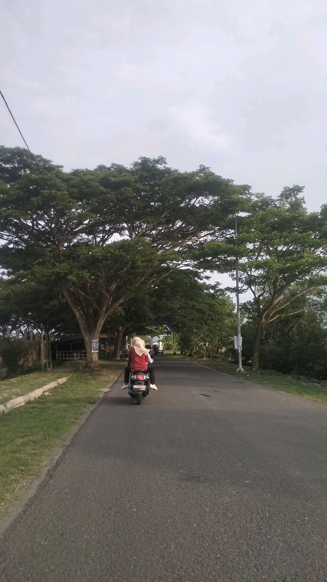 A scooter carries two individuals down a paved road flanked by lush green trees and grass. The person in the front is wearing a red top and a light-colored headscarf, while the person in the back is wearing a white top and dark pants. The scooter has a license plate that reads "BL 6958 KAH". The sky above is overcast with a pale gray hue. The scene appears to be in a semi-rural or park-like setting within Lhokseumawe, Indonesia, characterized by the large, mature trees and the presence of a road.